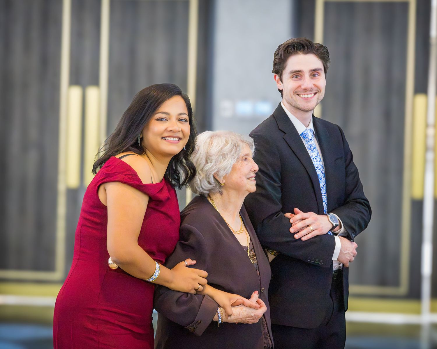 Two smiling young adults in formal attire posing with an elderly woman at an elegant celebration event.