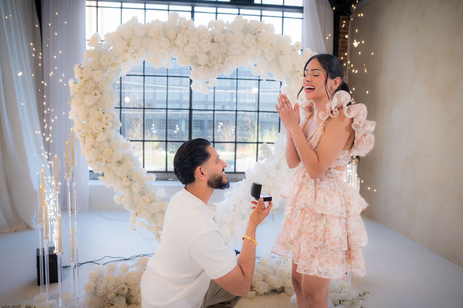 Man kneeling to propose to woman with ring box in front of a large white floral heart arch.