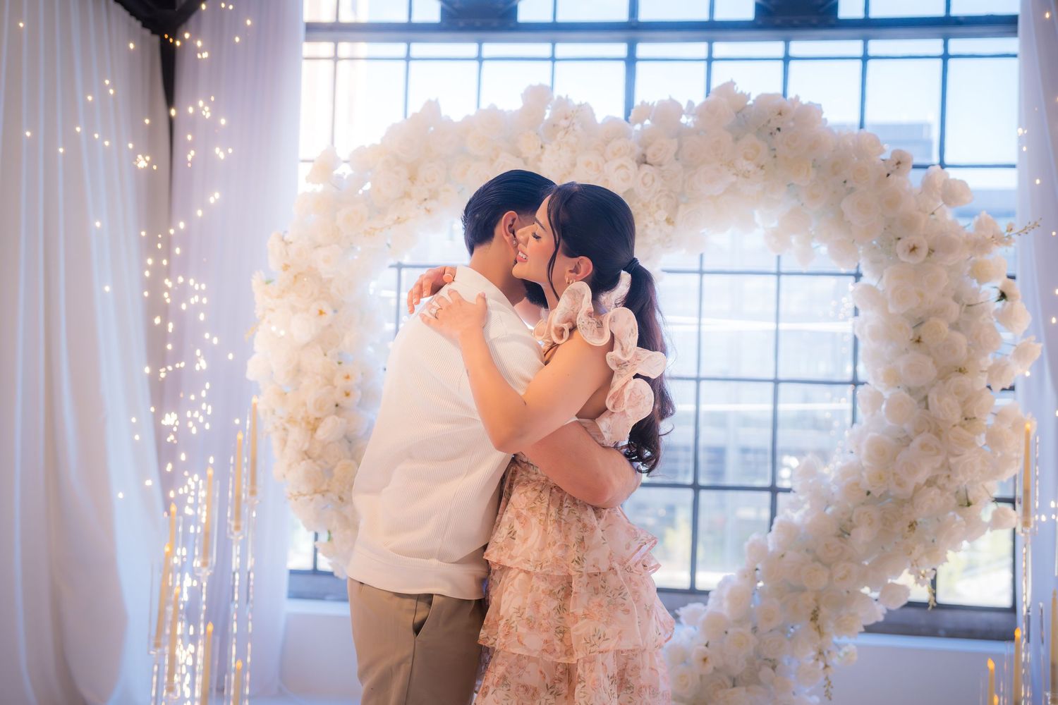 Couple sharing romantic kiss in front of white floral heart arch with twinkling lights.