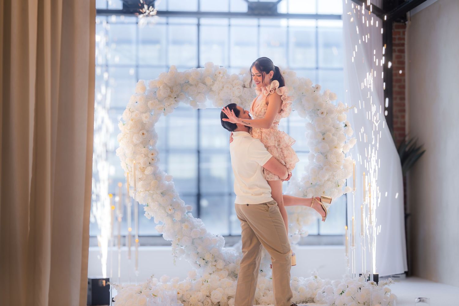 Man lifting his joyful fiancee in the air in front of a heart-shaped floral arch with fireworks.