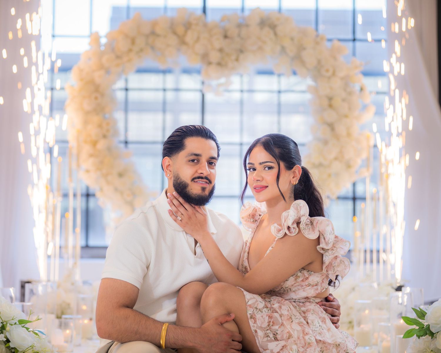 Newly engaged couple posing under white floral heart arch with sparklers behind them.