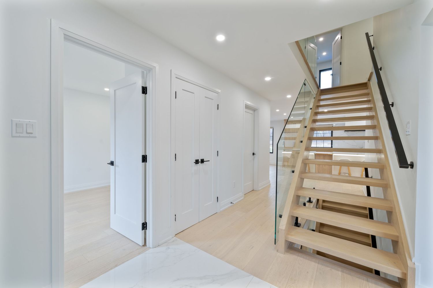 Modern white staircase with wooden treads and glass railing illuminated by recessed lighting in a bright hallway.