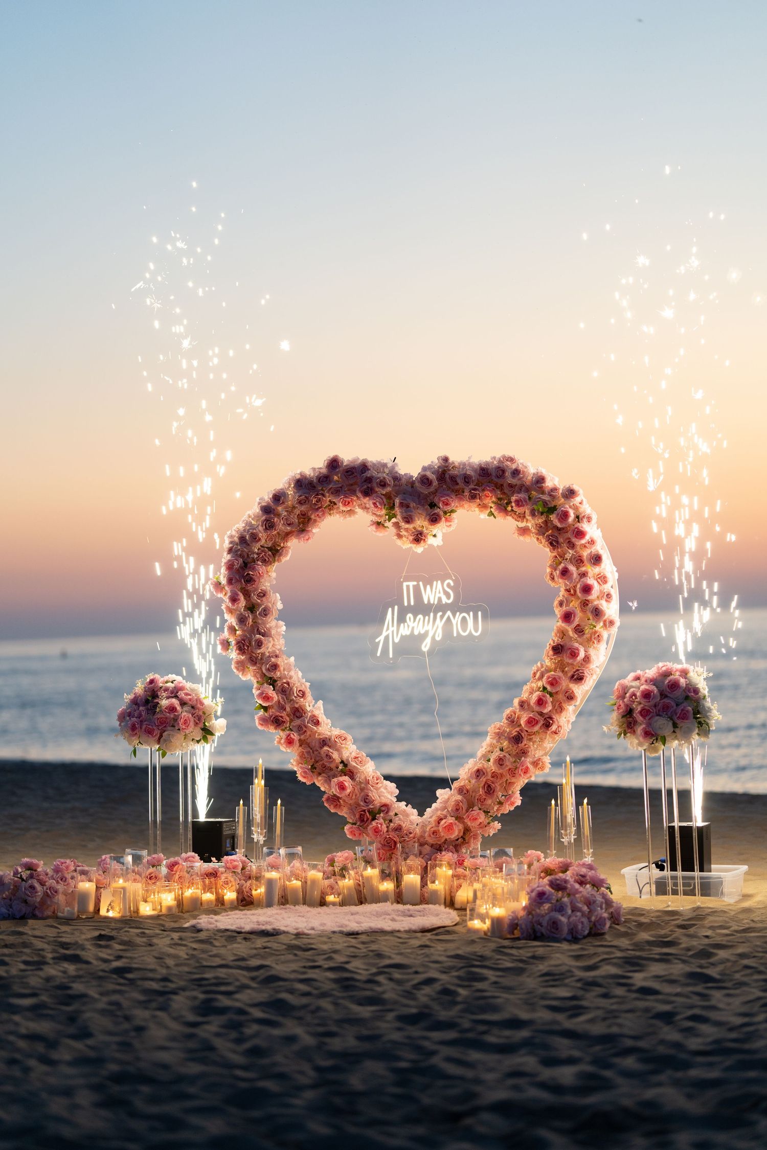 Heart-shaped flower arch with sparklers and candles on beach during sunset proposal.