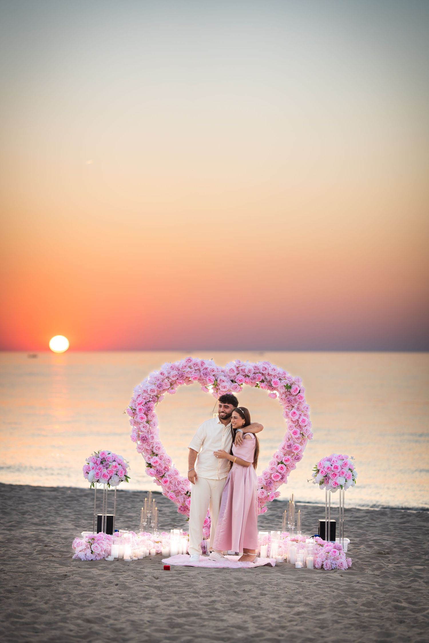 Romantic beach proposal setup with heart-shaped flower arch and sunset backdrop over ocean.