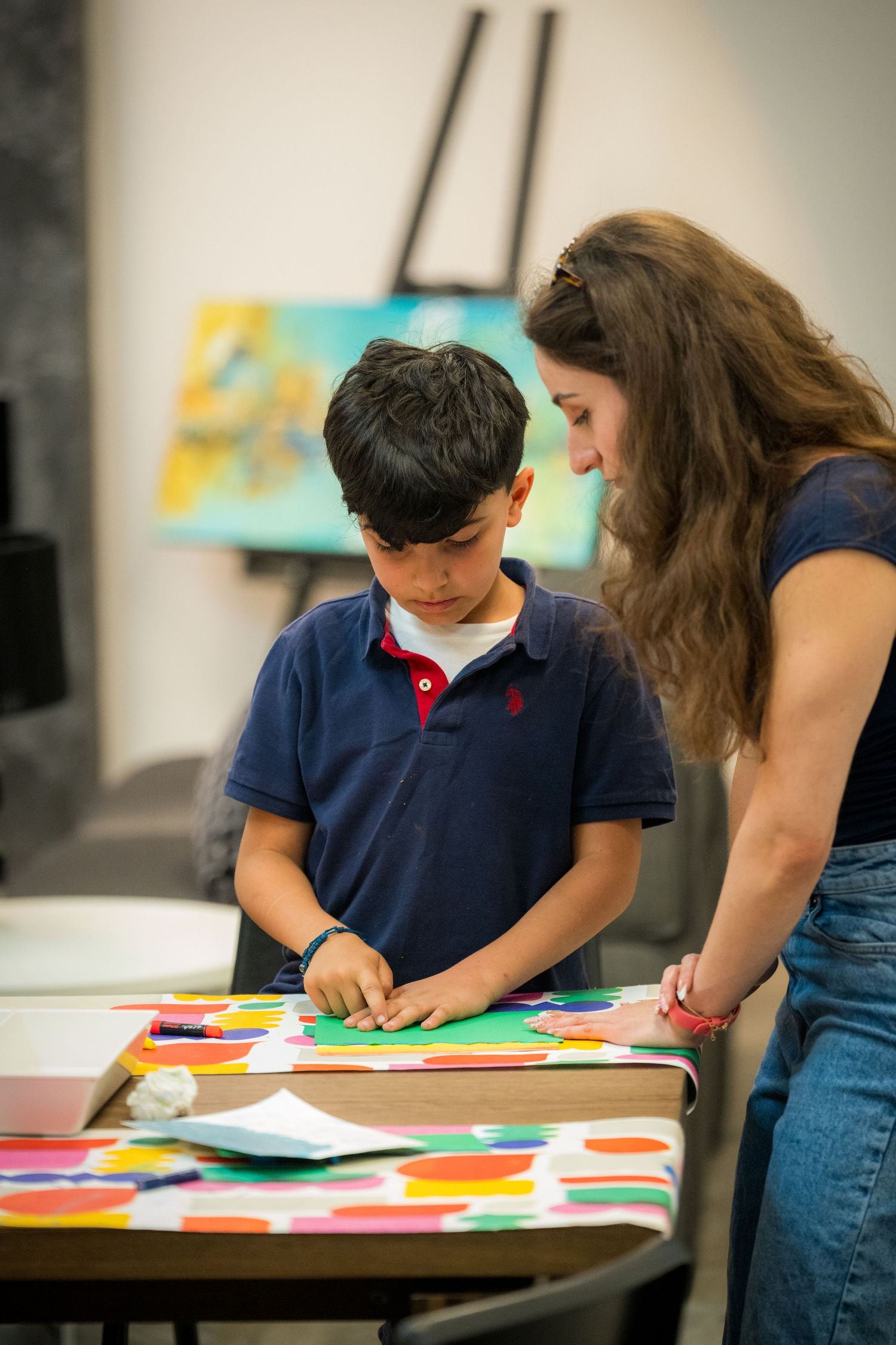 A woman helps a young boy with a colorful art project at a table in an art classroom.