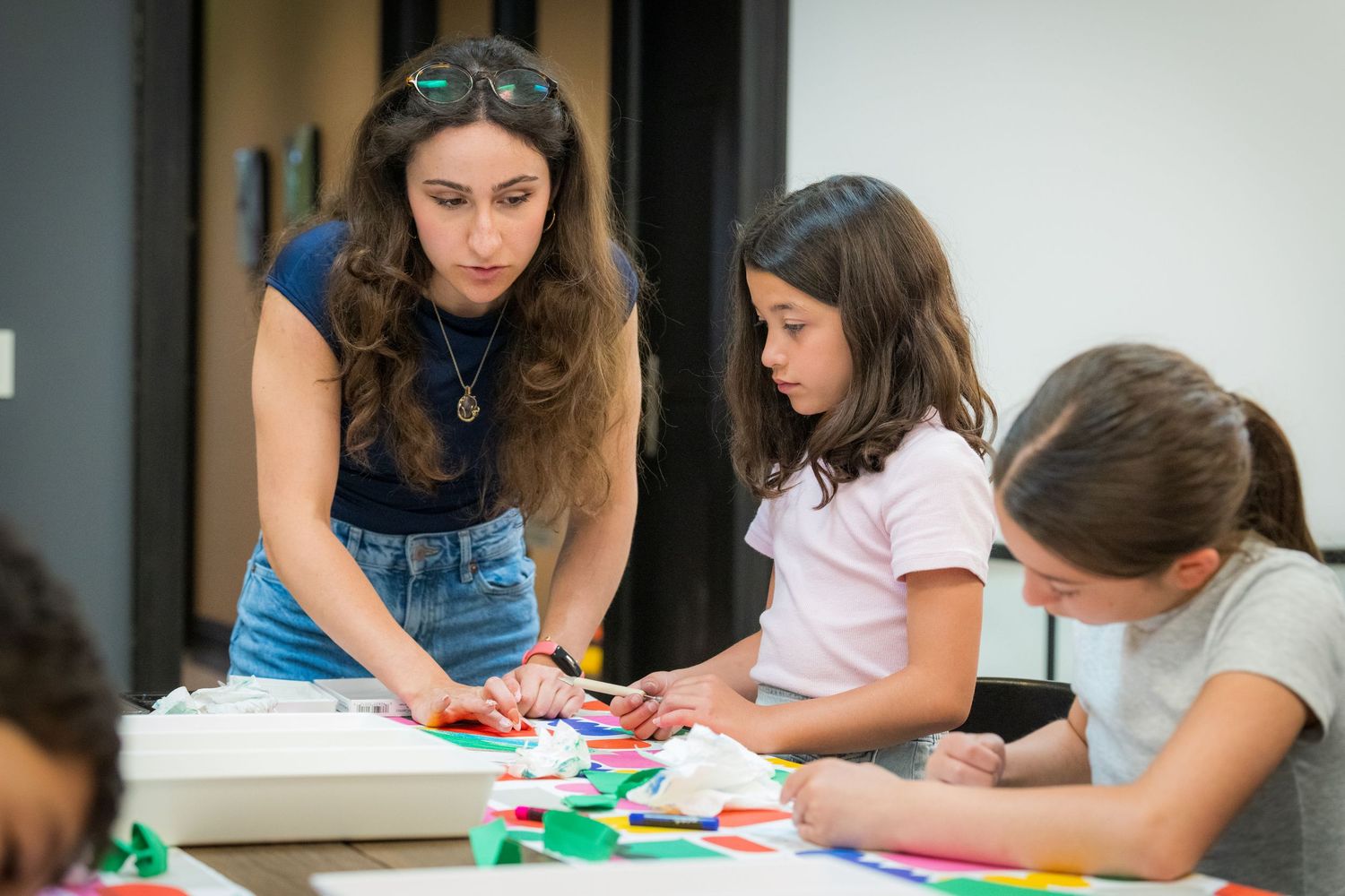 A teacher guides two girls in a colorful paper collage art workshop at a classroom table.