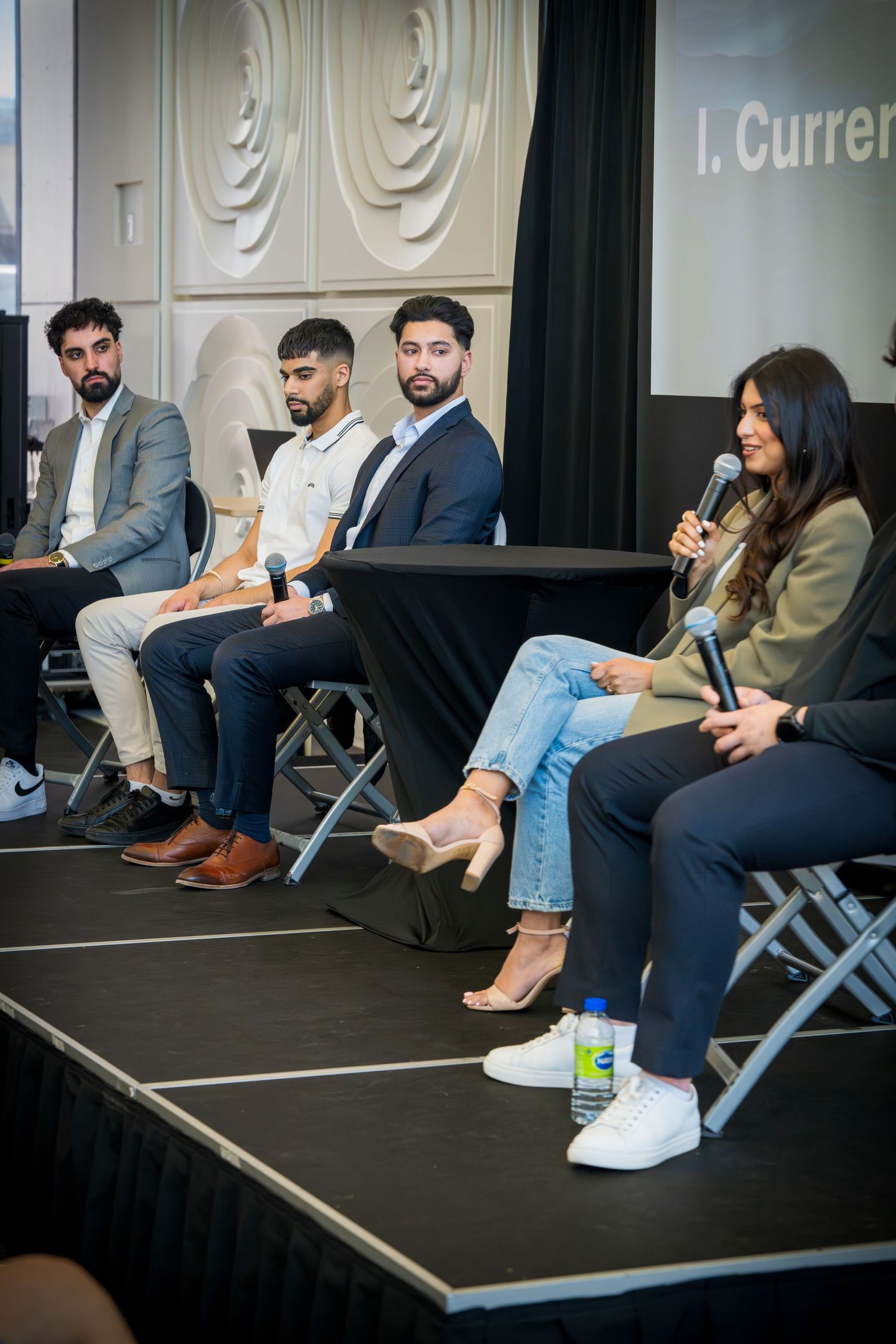 Conference panel discussion with speakers seated in row on raised platform with decorative wall backdrop.