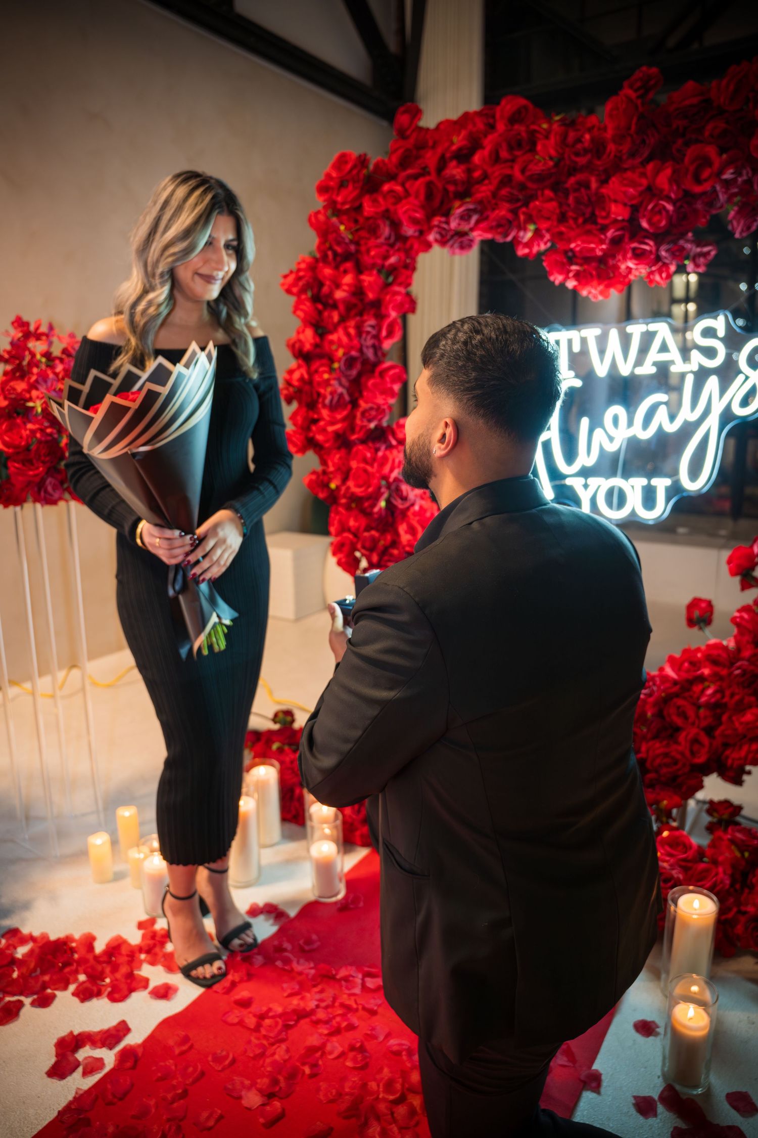 Romantic proposal in progress with stunning red rose wall and neon light decorations.
