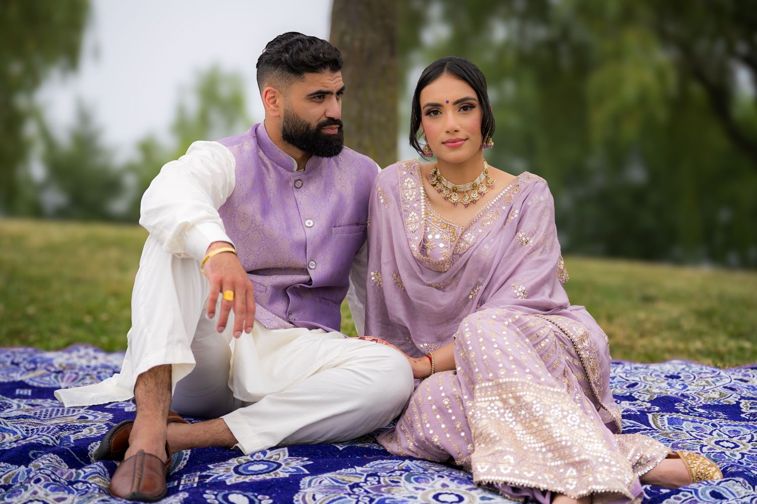 Elegant couple in matching purple and white outfits pose intimately on patterned blanket outdoors.