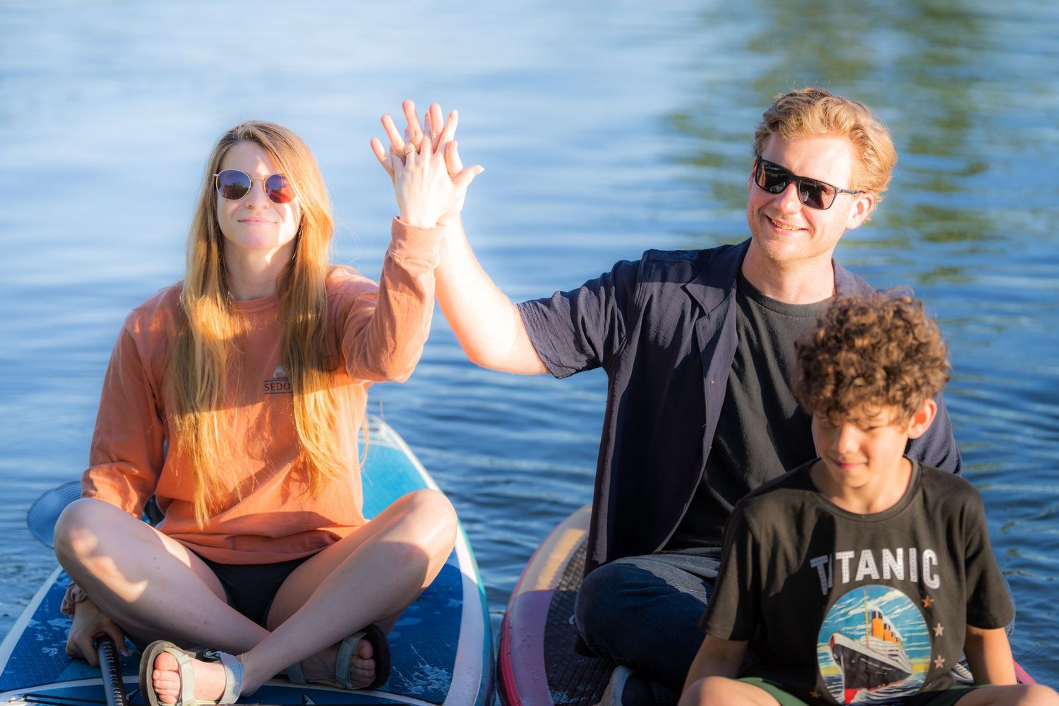 Engaged couple and friend celebrating on a paddleboard, waving on a sunny lake.