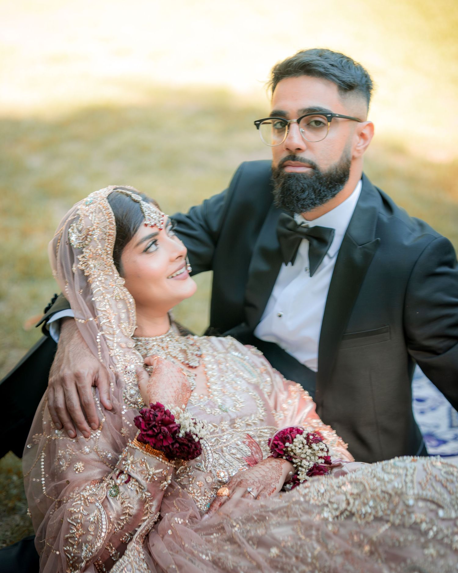 Couple in ornate South Asian wedding attire pose together on grass during golden hour.