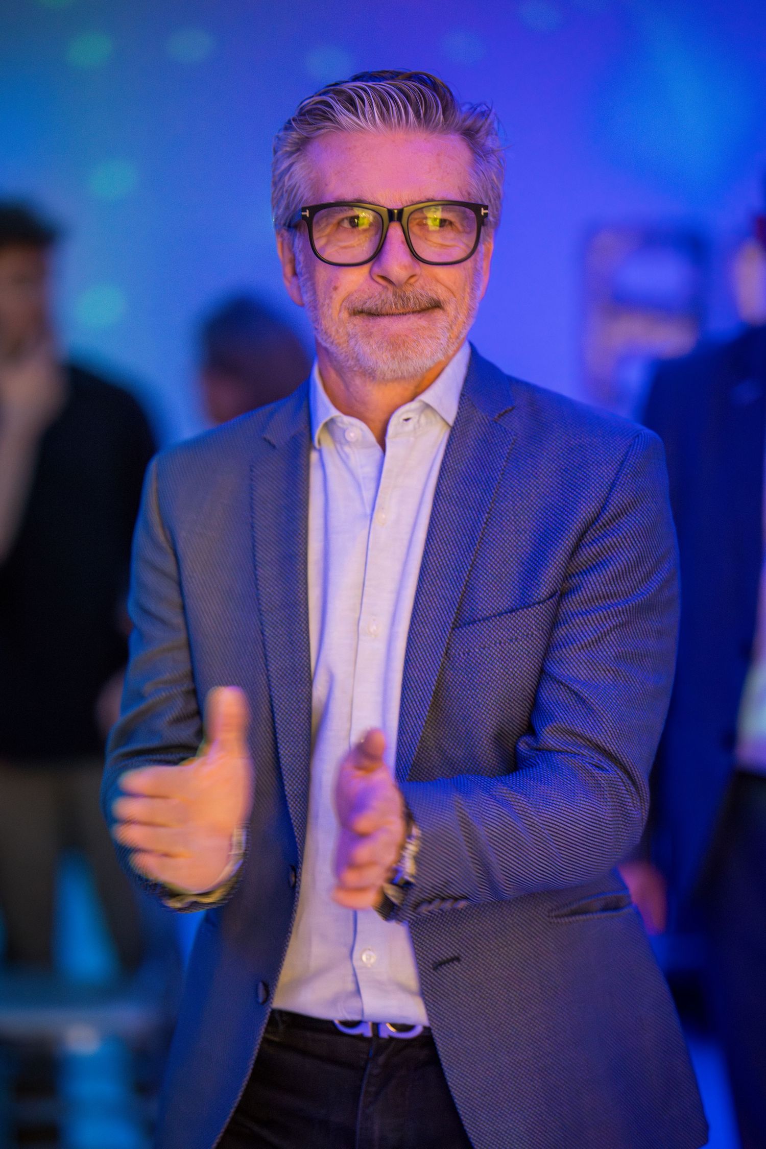 Person in blue suit and glasses gesturing expressively under dramatic blue lighting.