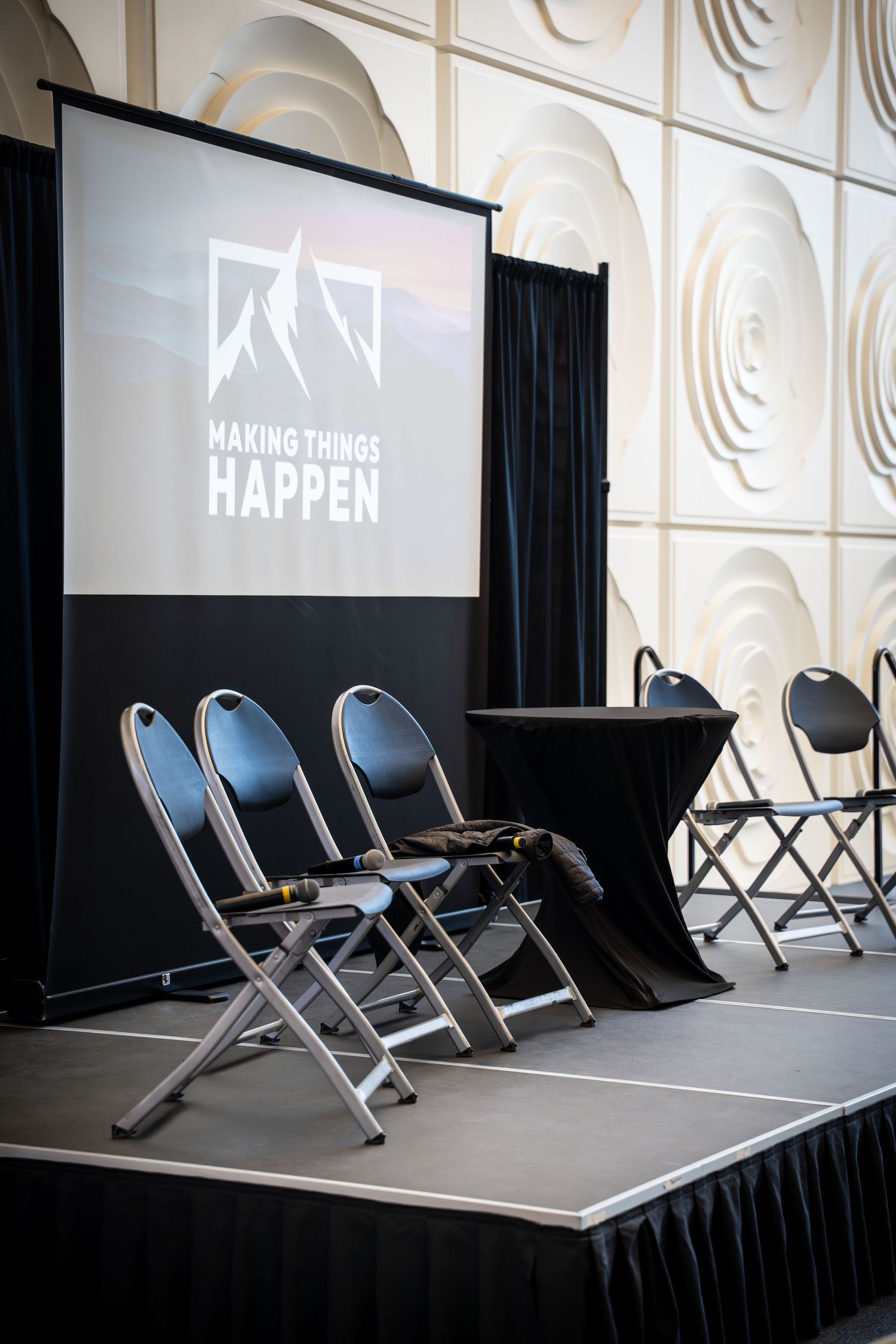 A stage setup with folding chairs and a projection screen displaying 'Making Things Happen'.