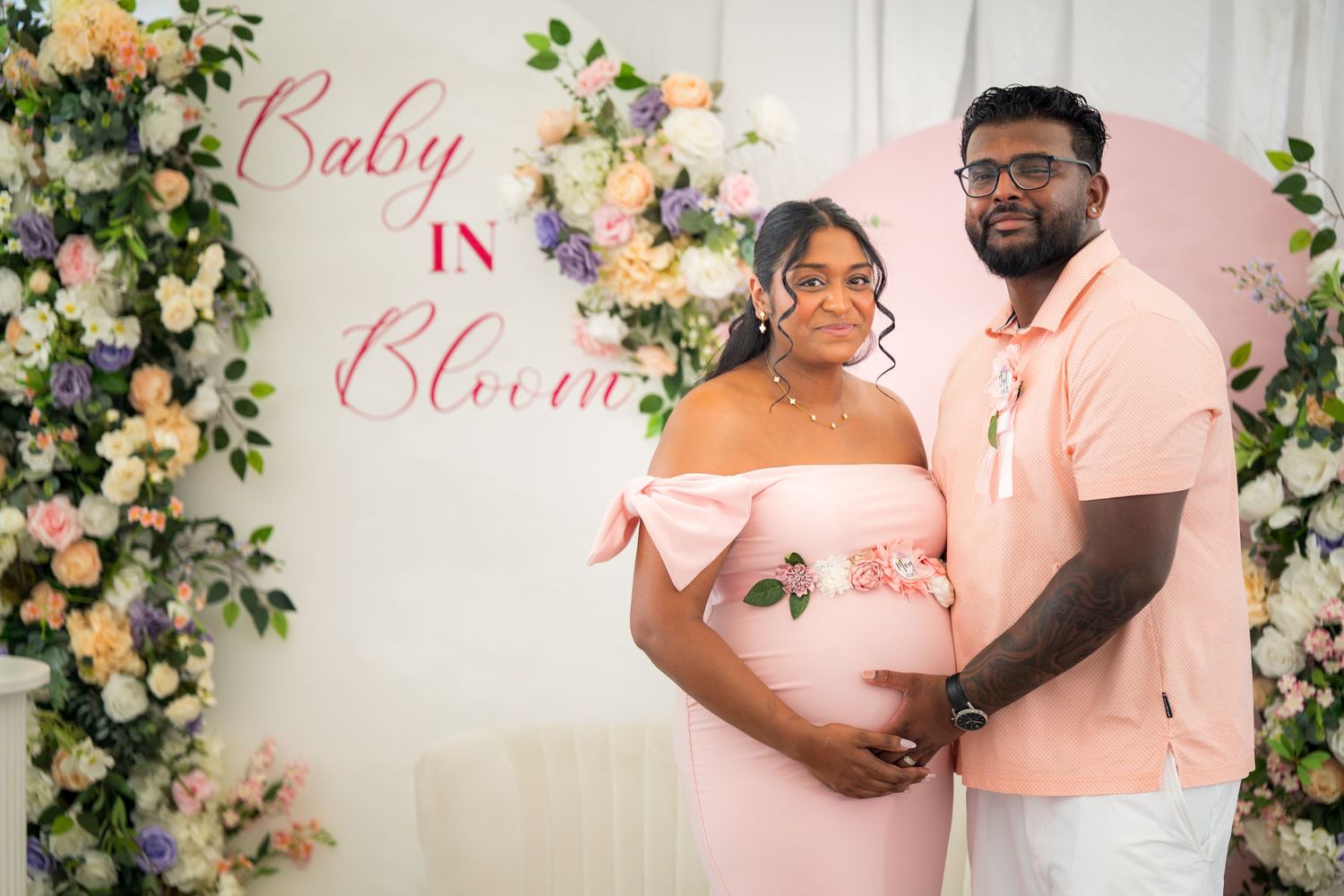 A couple stands together in matching pastel outfits against an elegant floral wall display.