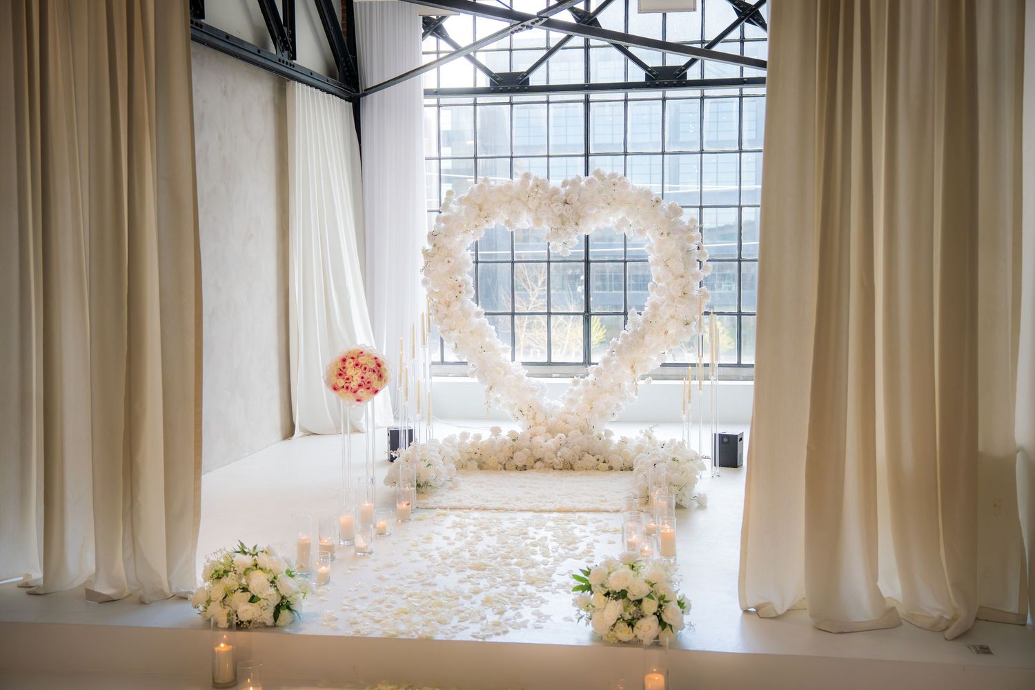 Elegant wedding proposal setup with a large white floral heart arch, rose petals on the floor, and candles in a bright venue.