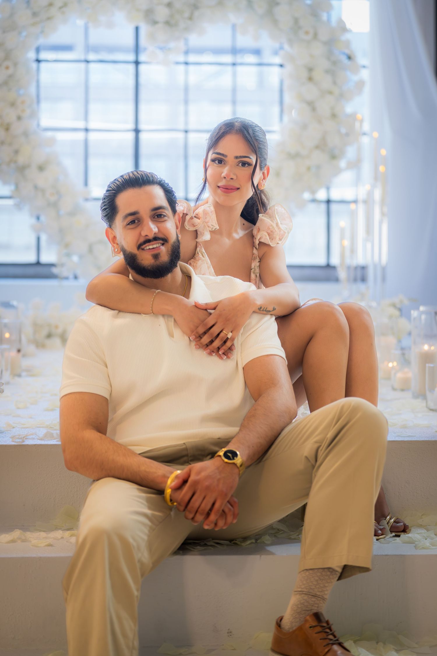 Couple posing at romantic white floral heart arch engagement setup, man seated with woman behind him.