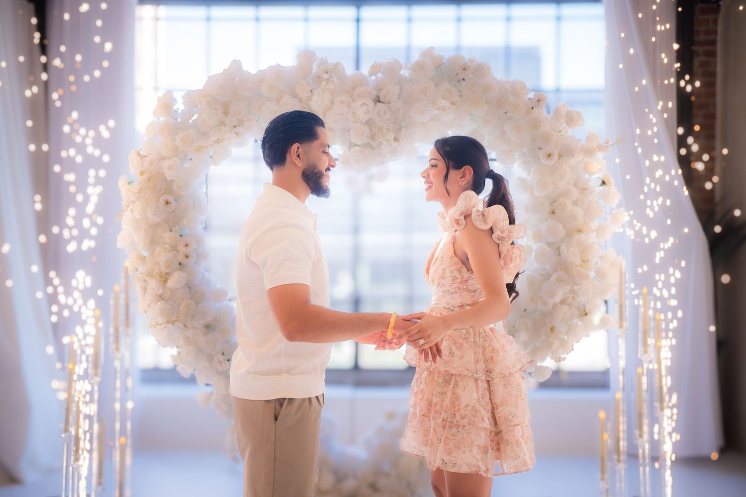 Couple holds hands and faces each other under a white floral heart arch with sparkling fairy lights.