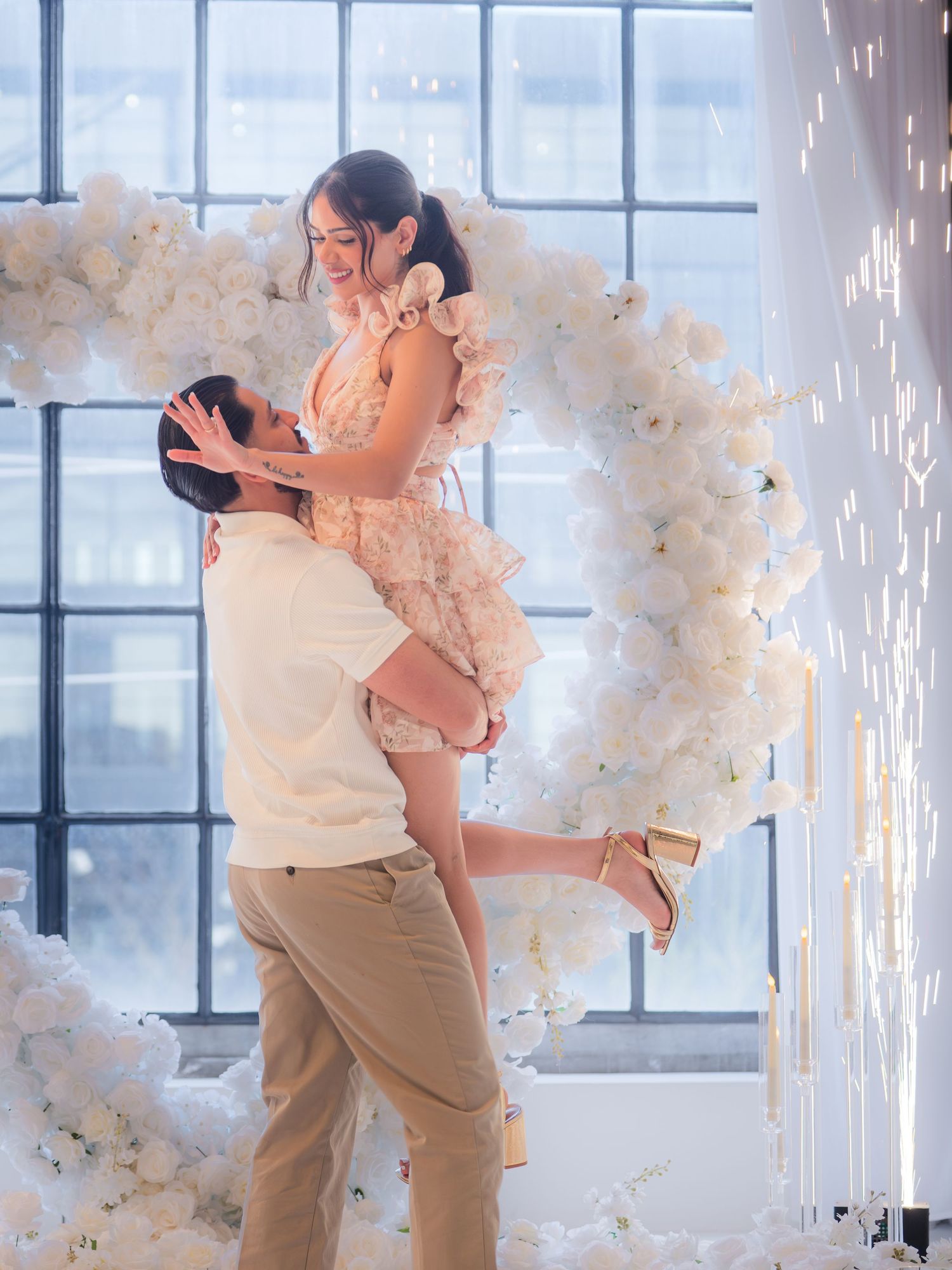 Man lifting smiling woman before a white flower heart arch with glittering sparkler displays.
