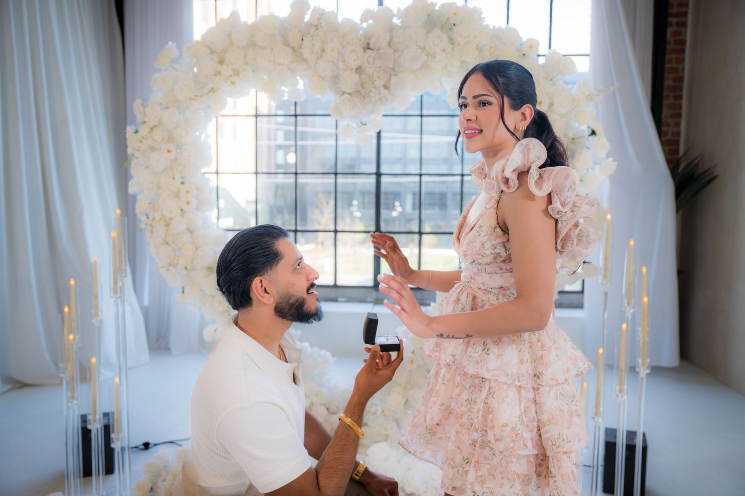 Man on one knee proposing to woman in floral dress with white heart floral arch backdrop.