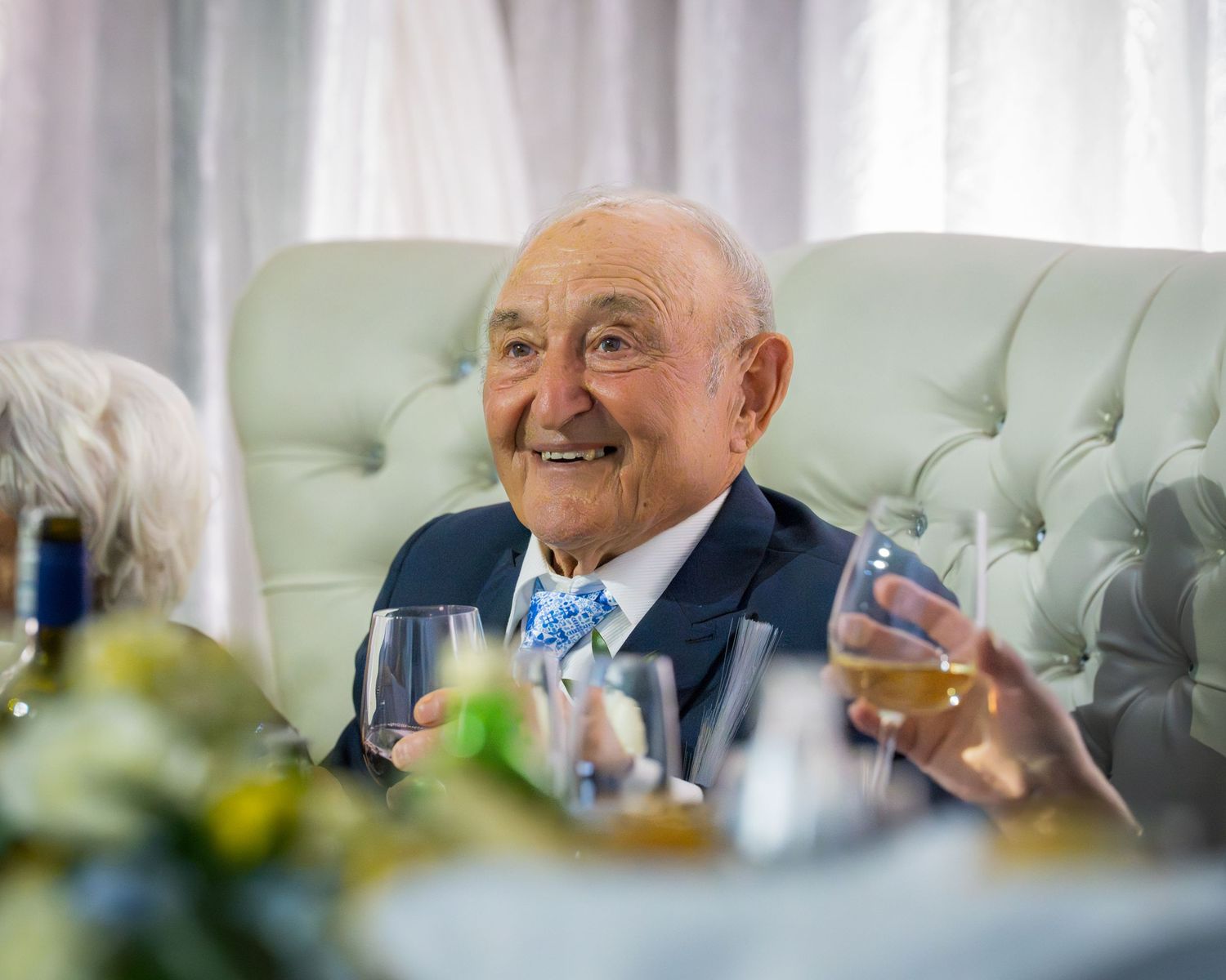 Happy elderly man in navy suit with blue bow tie smiling at anniversary celebration dinner table.