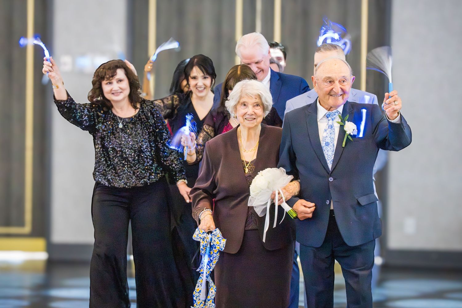 Elderly couple leading a joyful procession waving ribbon wands at a formal anniversary celebration event.