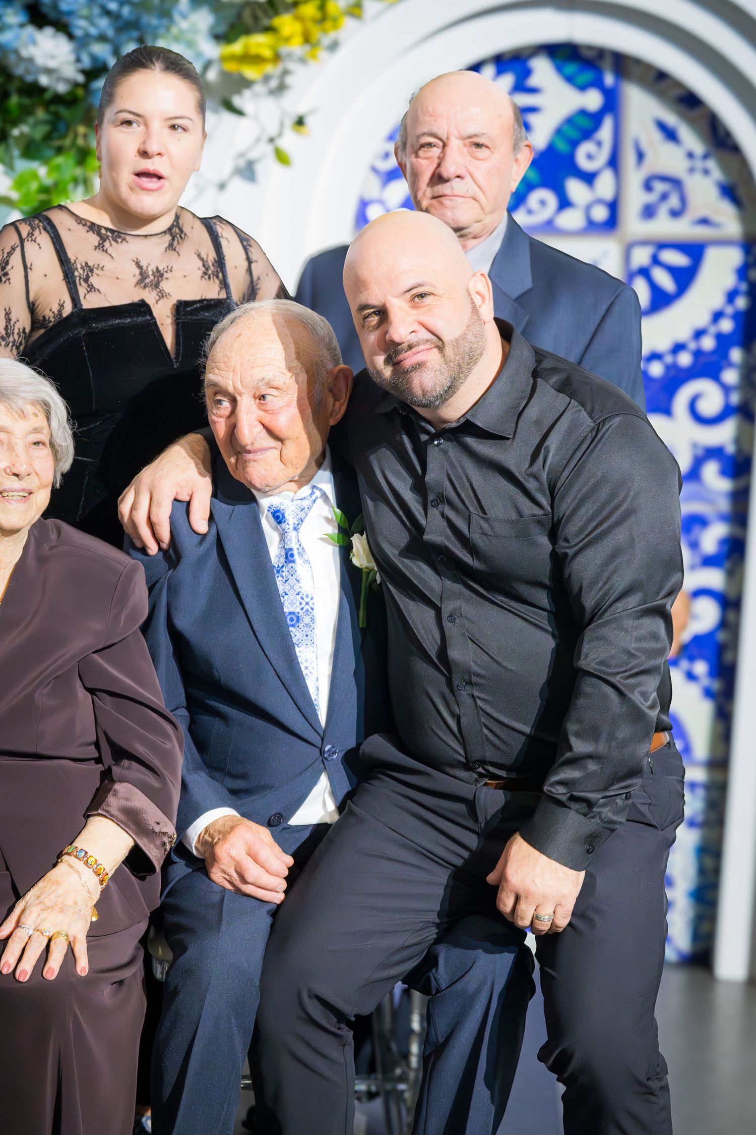 Bald man in black shirt embraces elderly man in suit at anniversary party with family members.