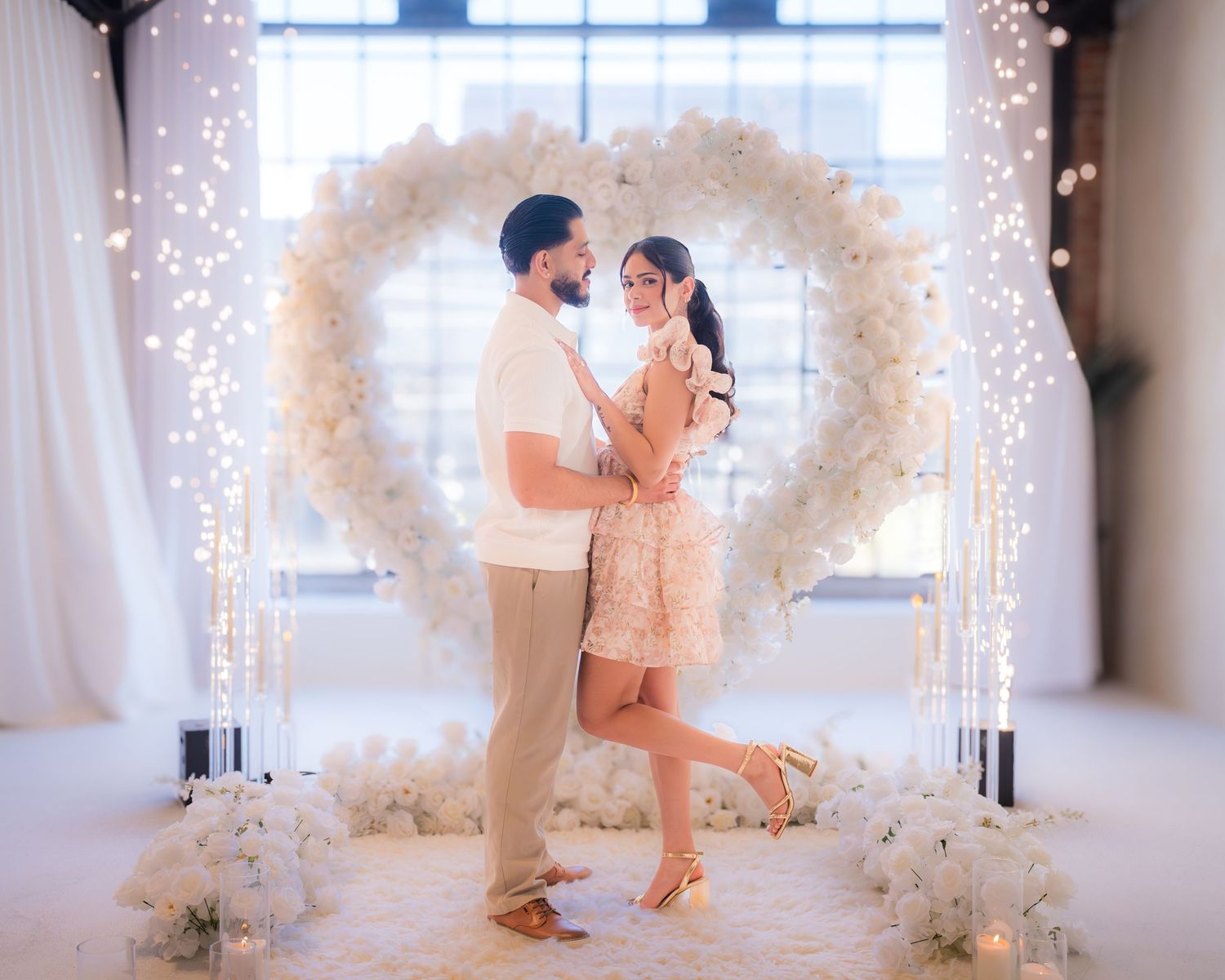 Couple embracing in front of a white floral heart arch with fairy lights at a romantic engagement setup.