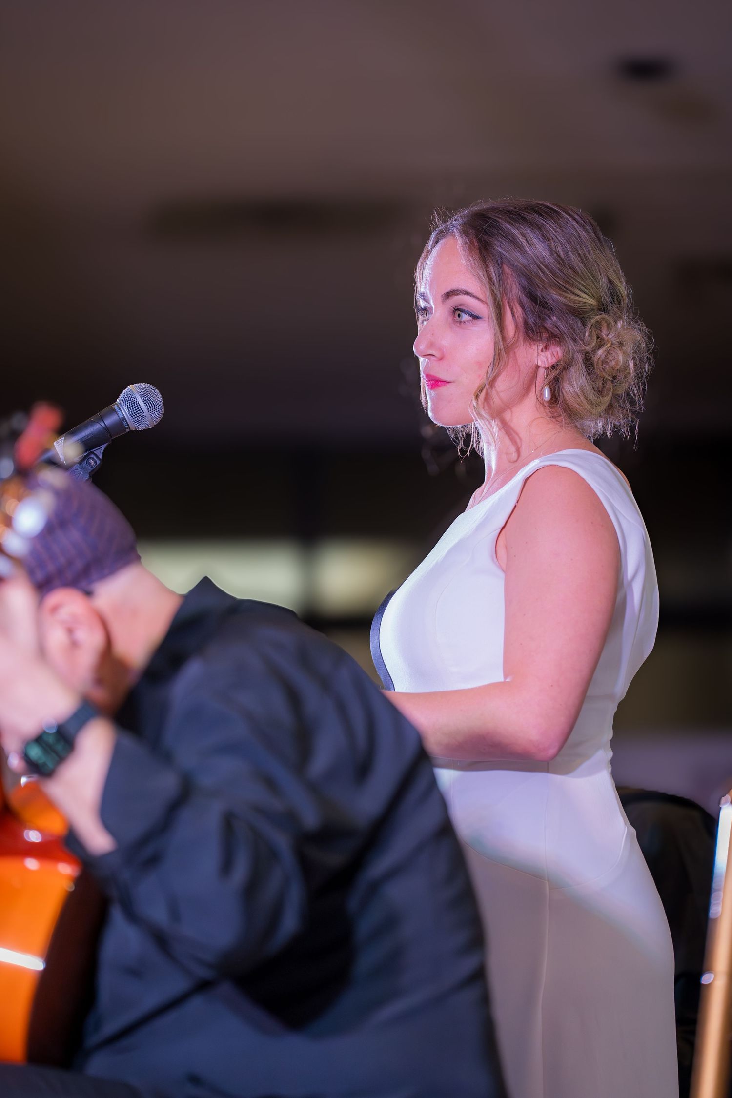 Elegant female singer in white dress stands by microphone while guitarist performs in foreground.