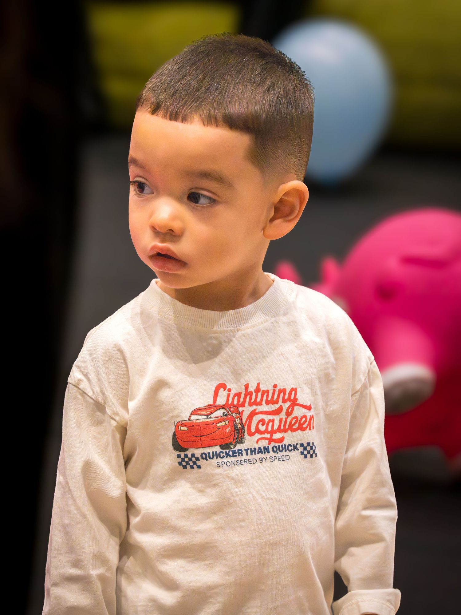 Young toddler boy wearing a Lightning McQueen white long sleeve shirt standing at an indoor play area.