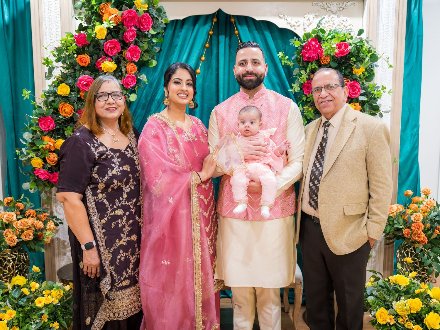 Smiling family group with baby in arms standing before colorful flower arch at ceremony.