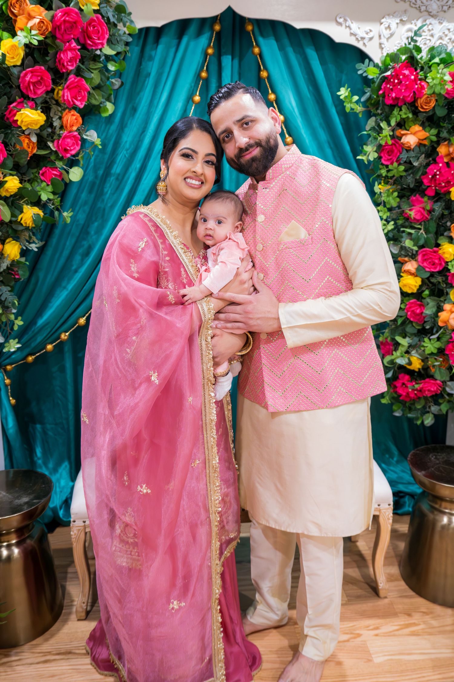 Indian family portrait with parents in pink and cream traditional attire holding newborn baby at floral decorated ceremony.