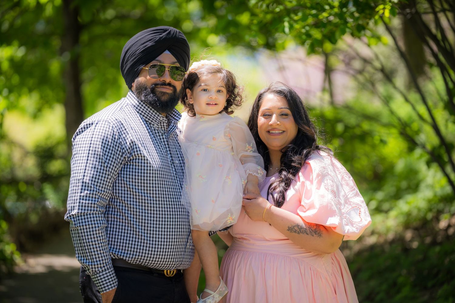 Family of three with father in turban, mother in pink dress, holding young child outdoors.