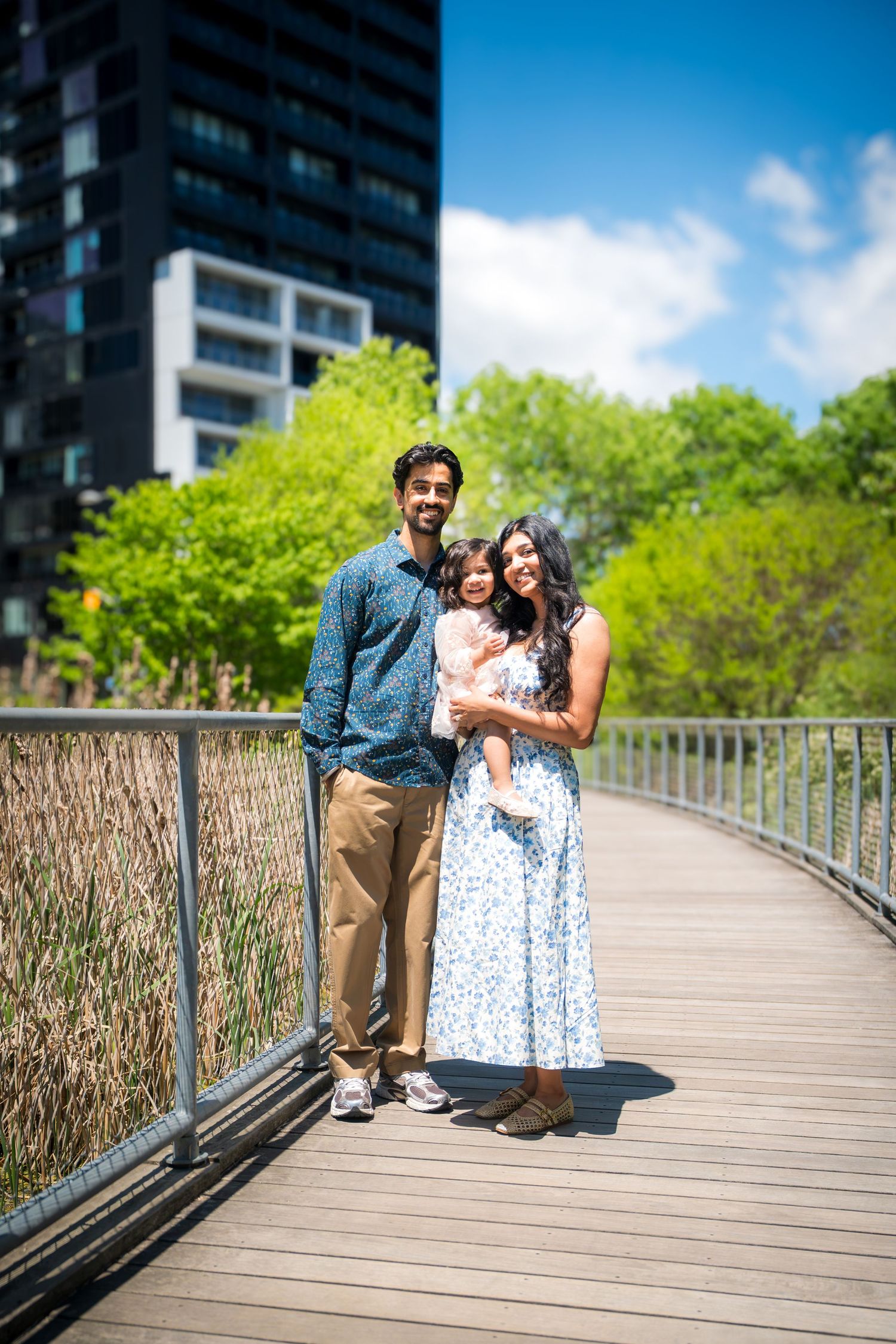 A family of three poses on a wooden boardwalk surrounded by lush green trees and a modern building on a sunny day.