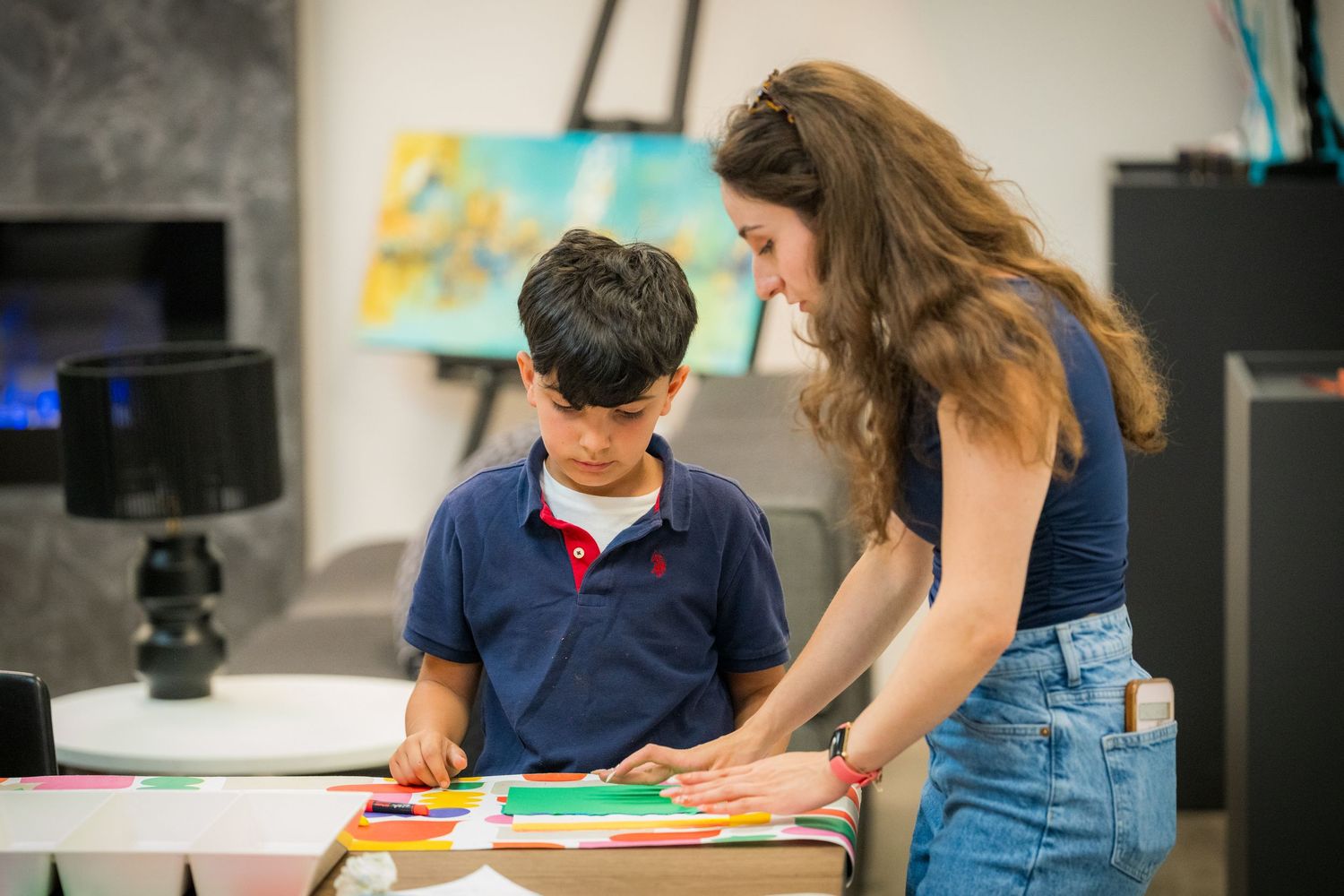 Female art teacher assists young boy with colorful paper craft project at creative children's workshop.
