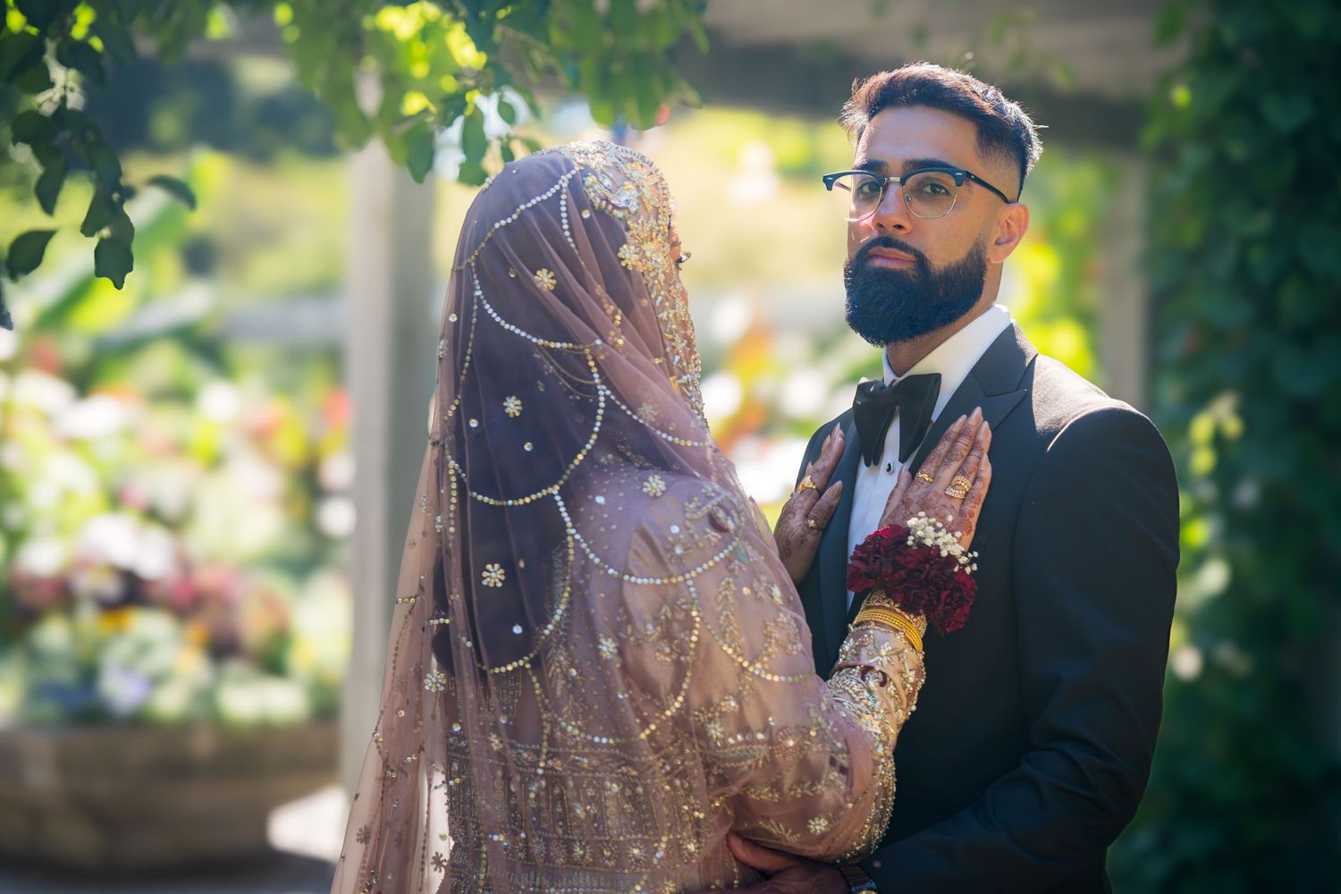 A couple shares a tender moment in a garden setting during their wedding celebration, with soft sunlight filtering through trees.