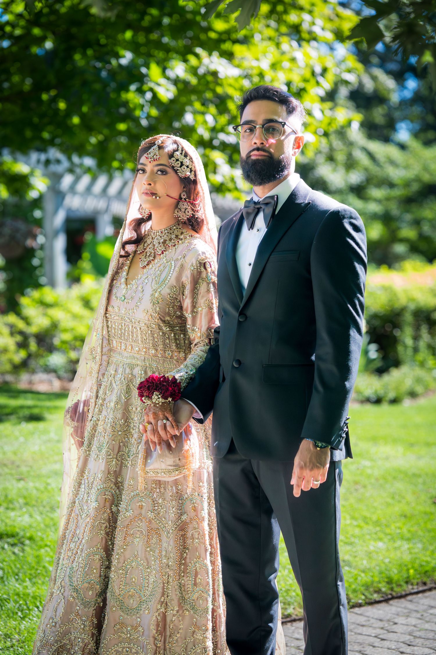 Couple in traditional South Asian wedding attire pose together outdoors with lush greenery in background.