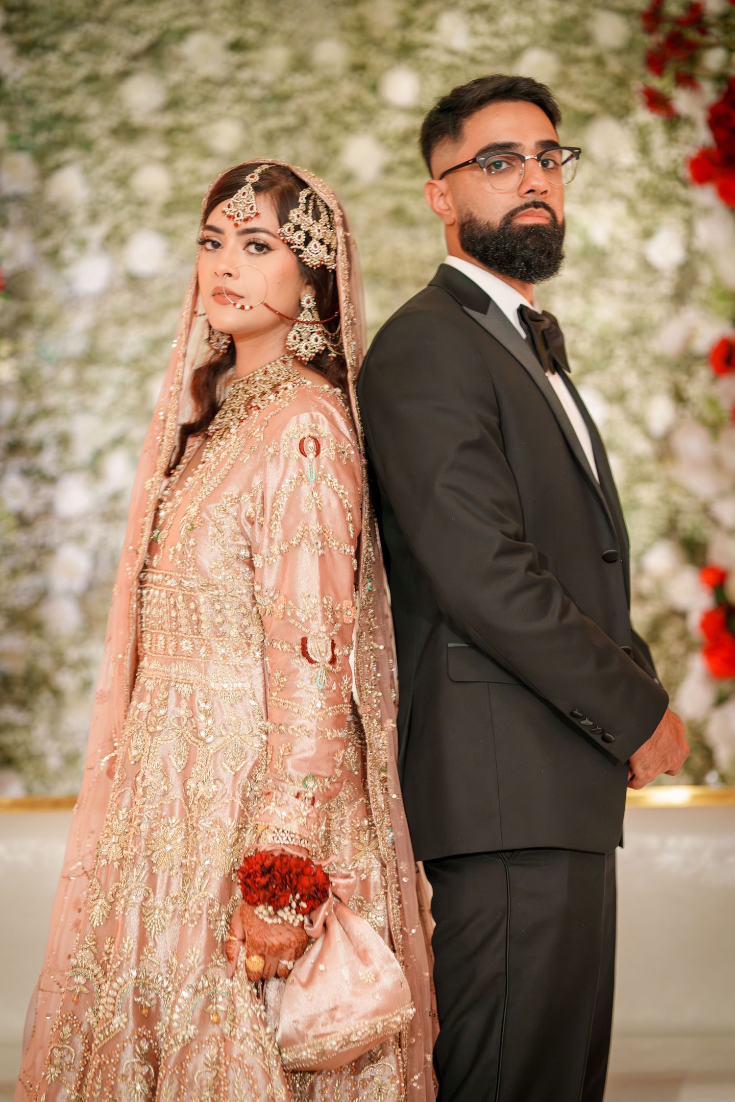Wedding couple in traditional attire pose against floral backdrop with red roses.