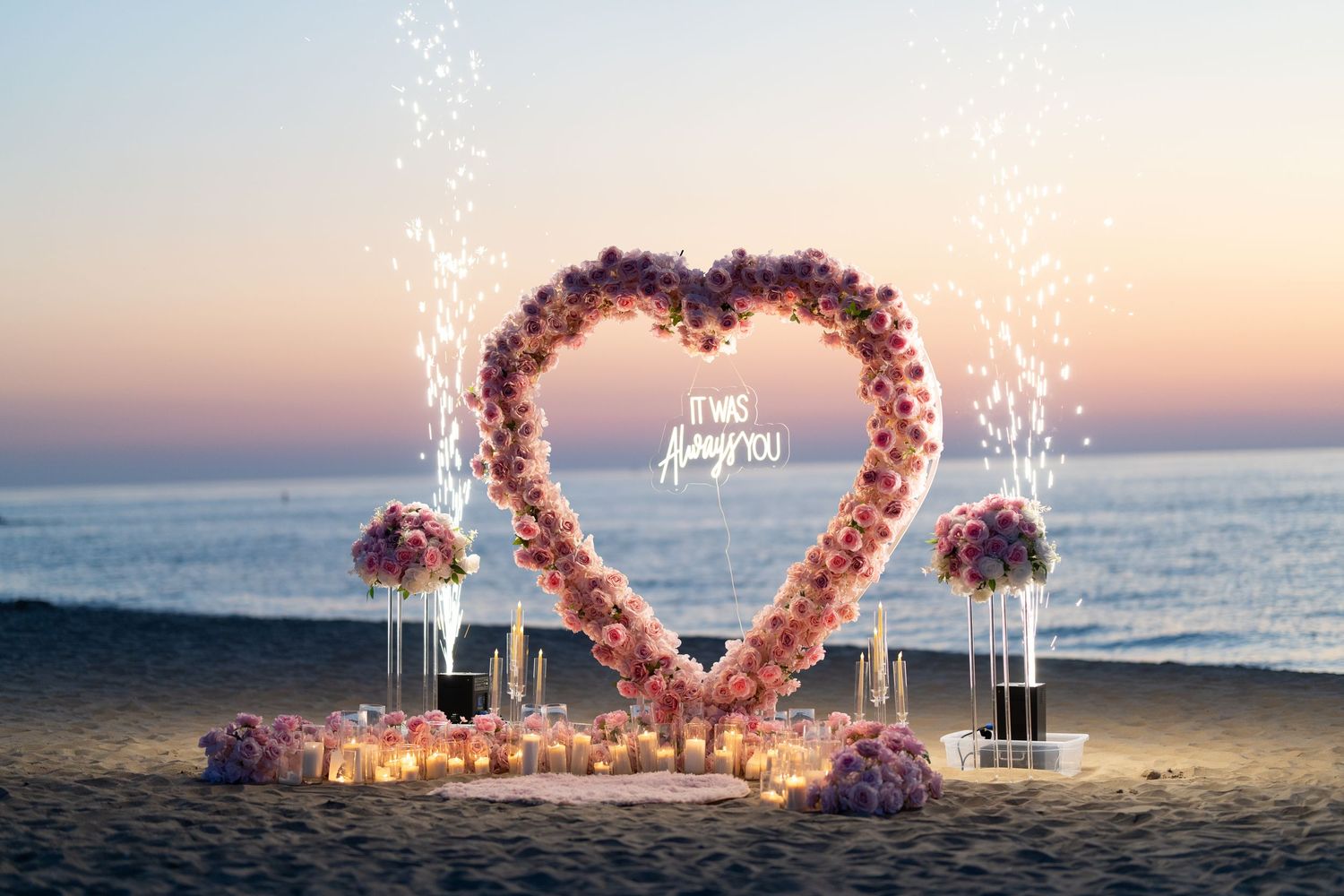 Pink floral heart arch with sparkling lights on beach at sunset for romantic proposal.