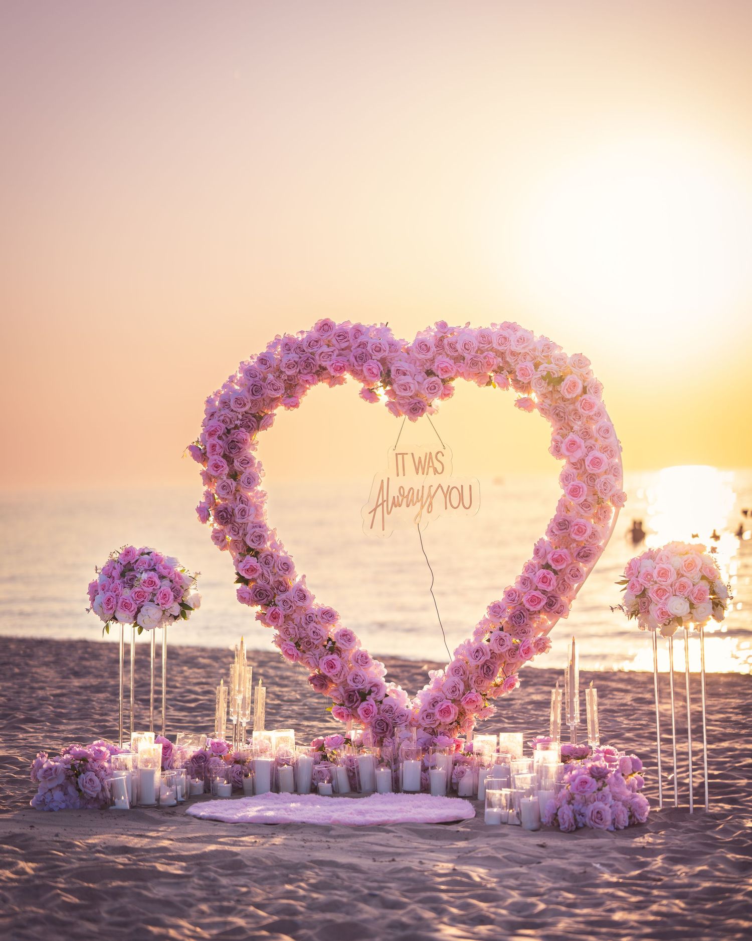Romantic beach proposal setup with purple flower heart arch and candles at sunset.