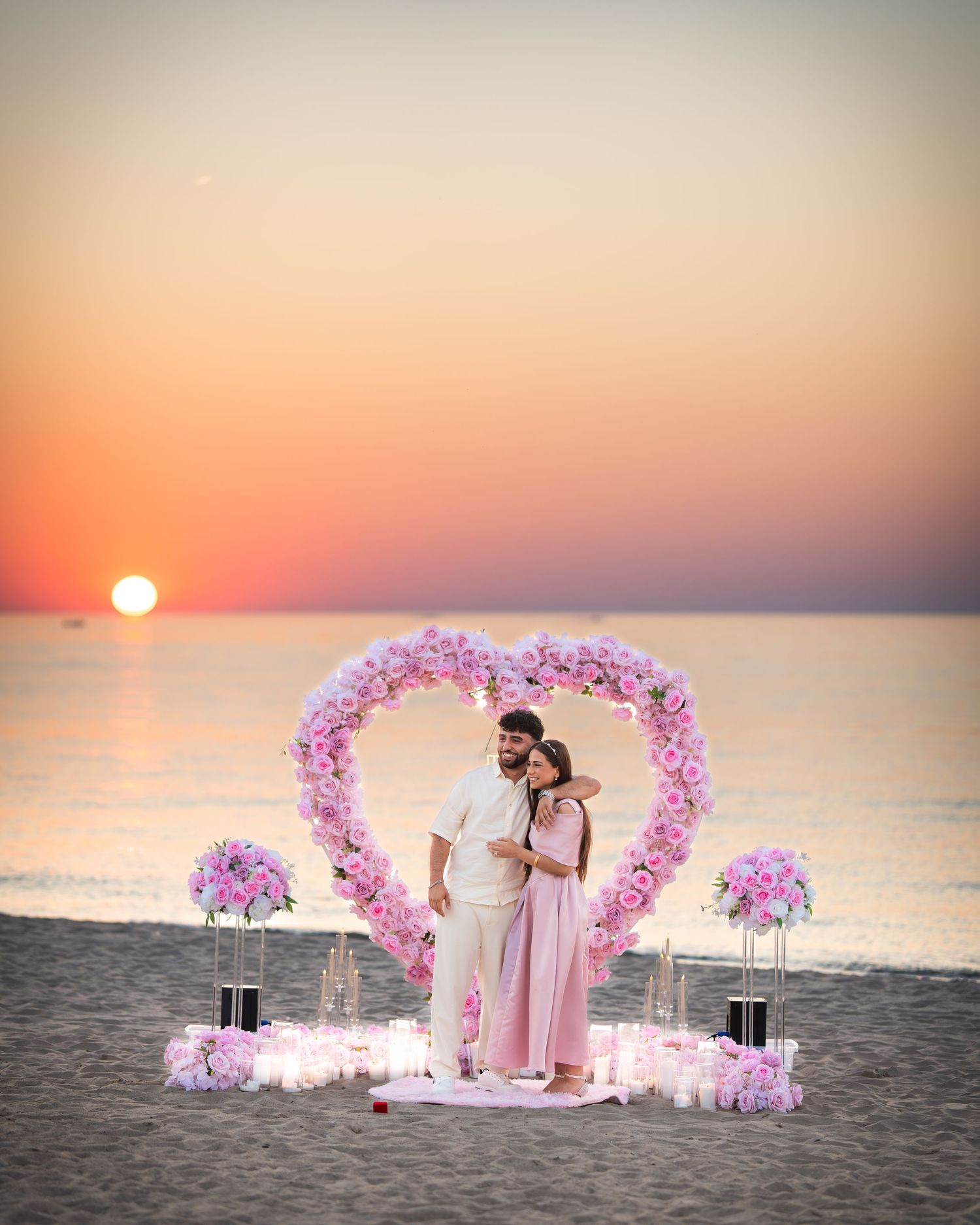 A romantic beach scene with a heart-shaped flower arch and pink decor at sunset over the ocean.