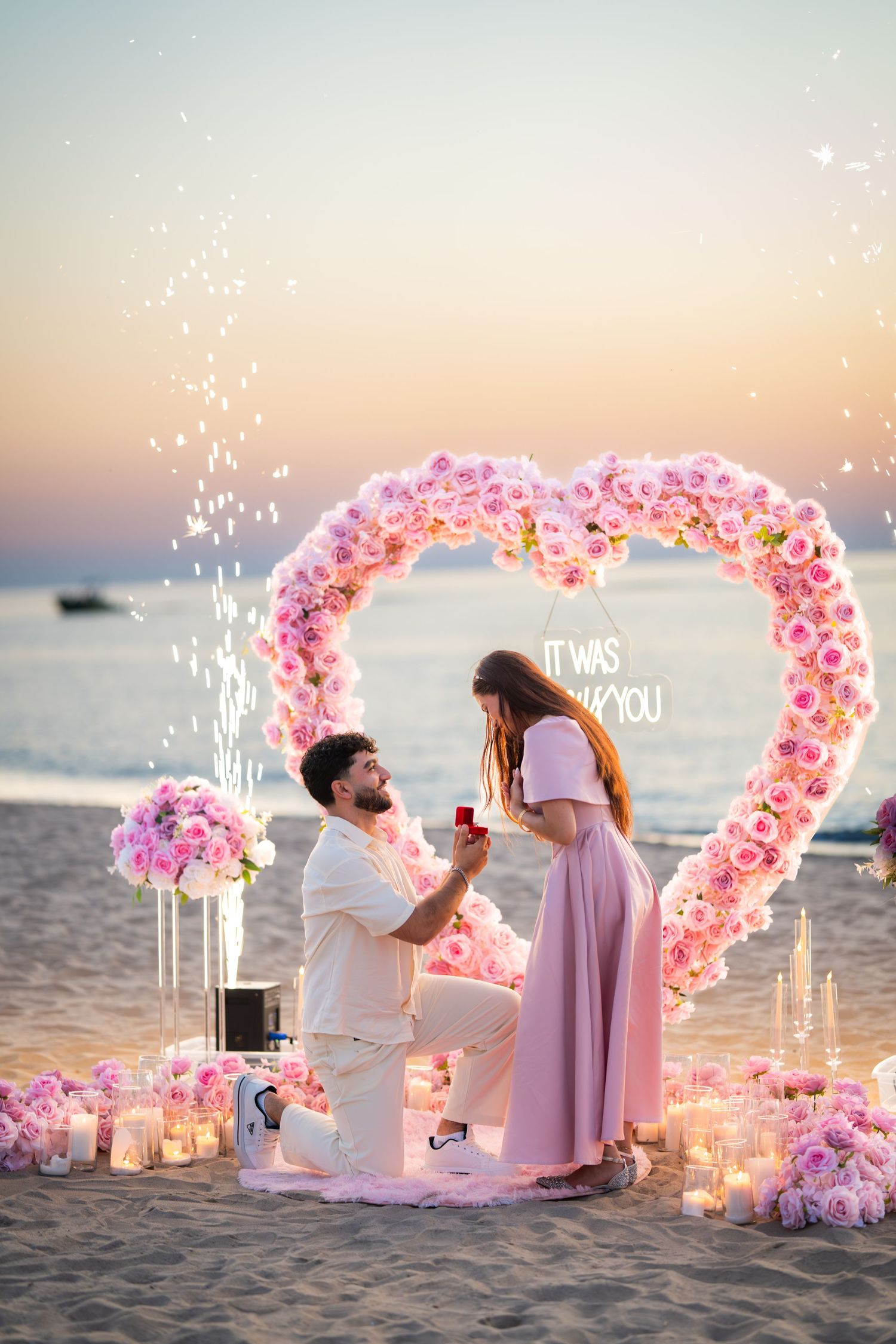 Romantic beach proposal setup with pink heart-shaped flower arch and candles at sunset.