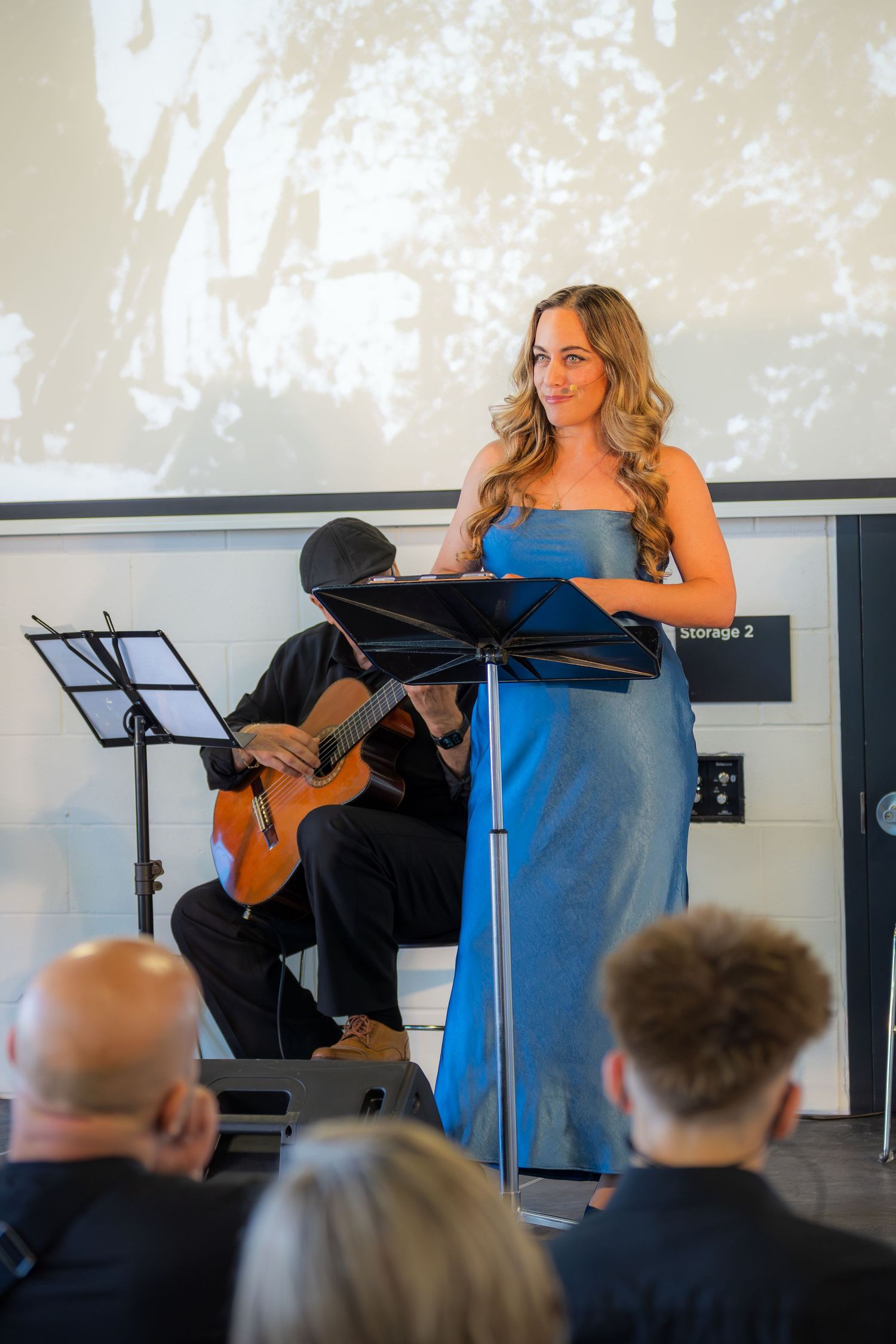 Woman in blue gown singing at music stand with guitarist accompanist performing live classical concert.