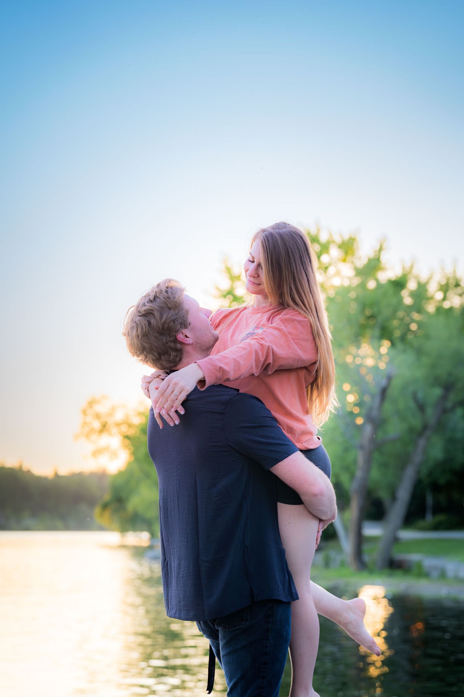 Man holding a woman in his arms near a sunlit lake at dusk, both smiling at each other.