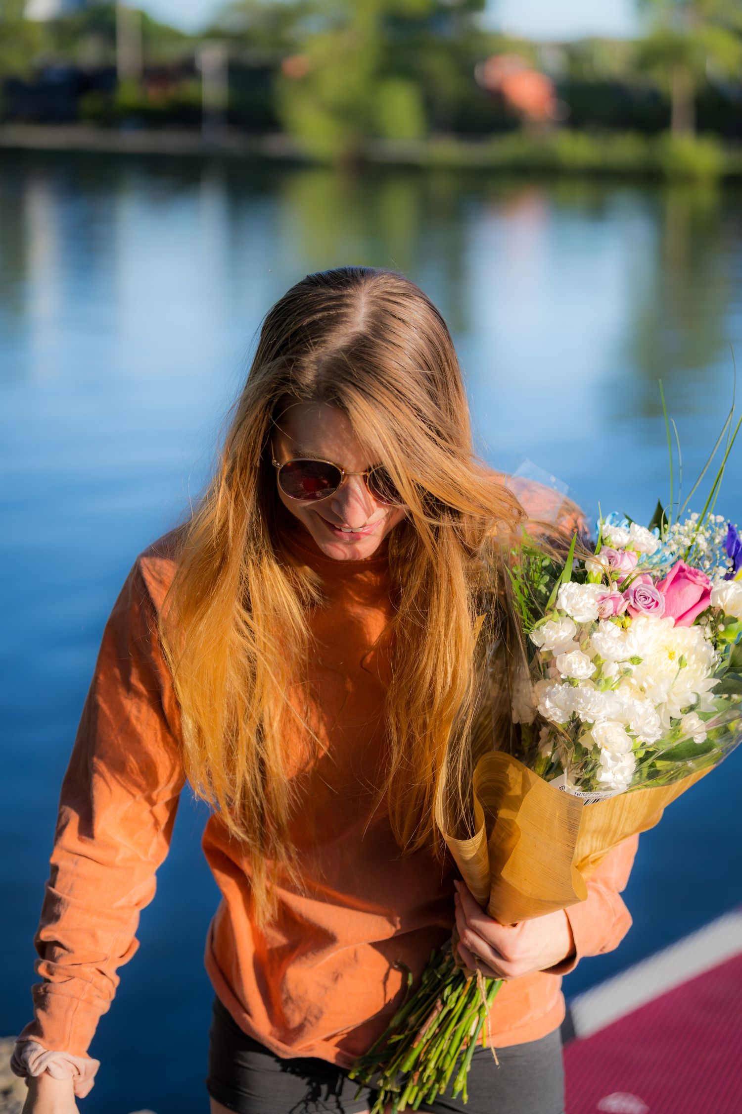 Woman in orange top holding a beautiful bouquet of flowers beside a calm blue lake.