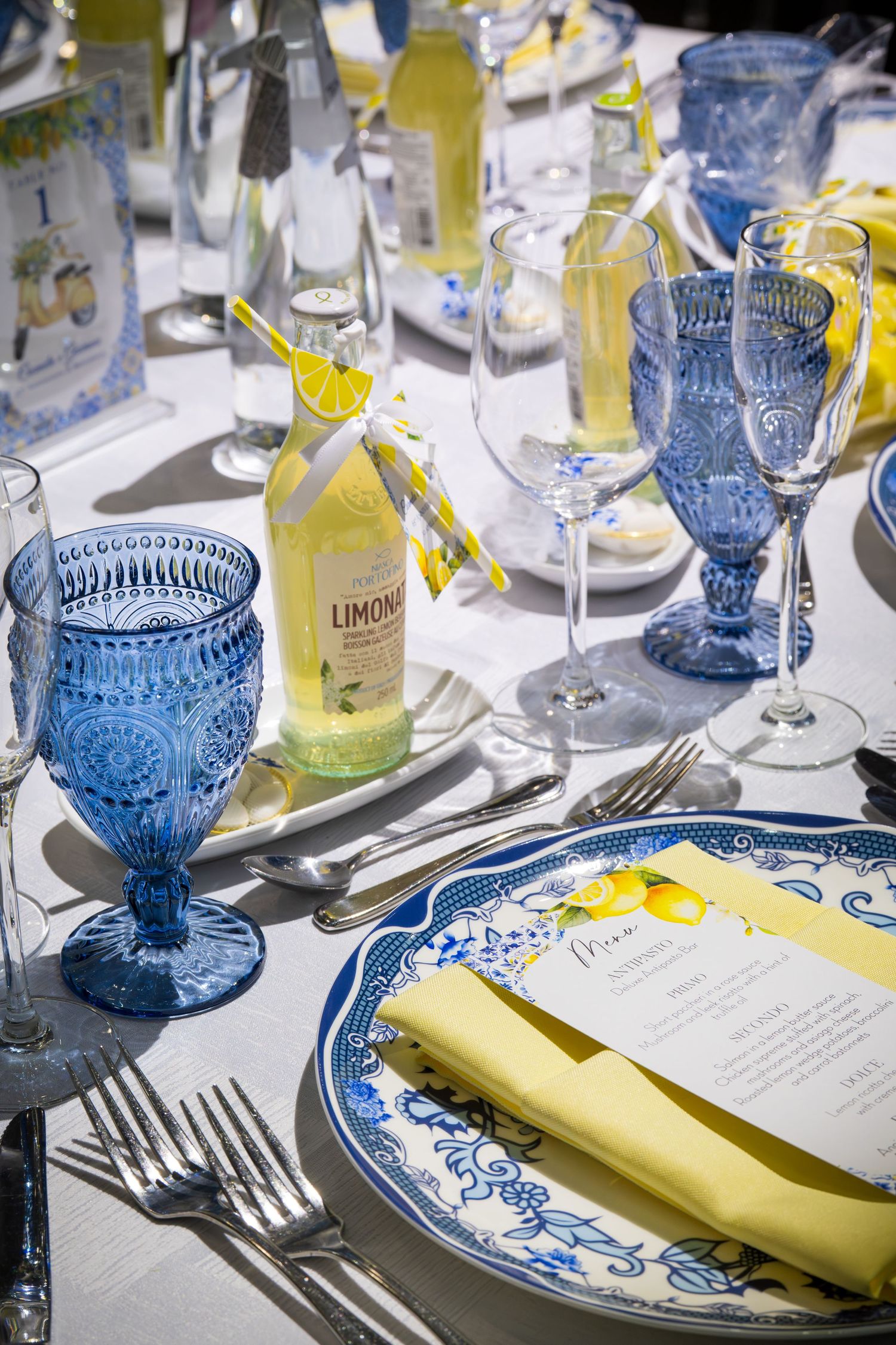 Elegant Italian-themed table setting with blue glassware, Limoncello bottle, blue and white plates, and yellow napkins.