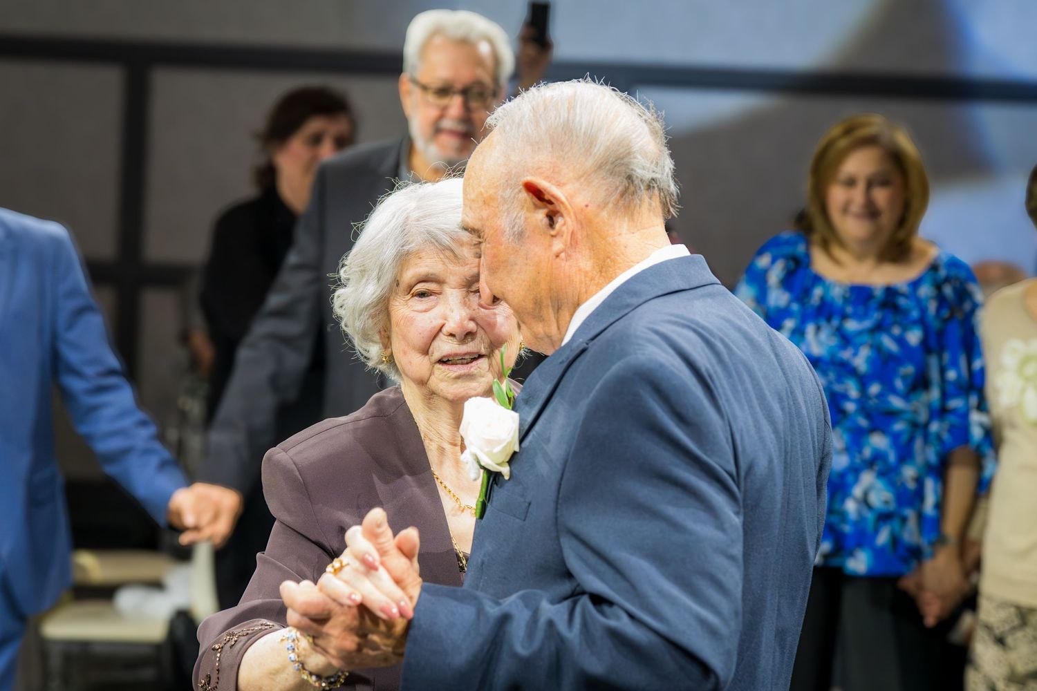 Elderly couple dancing together at an anniversary party surrounded by smiling guests in formal attire.