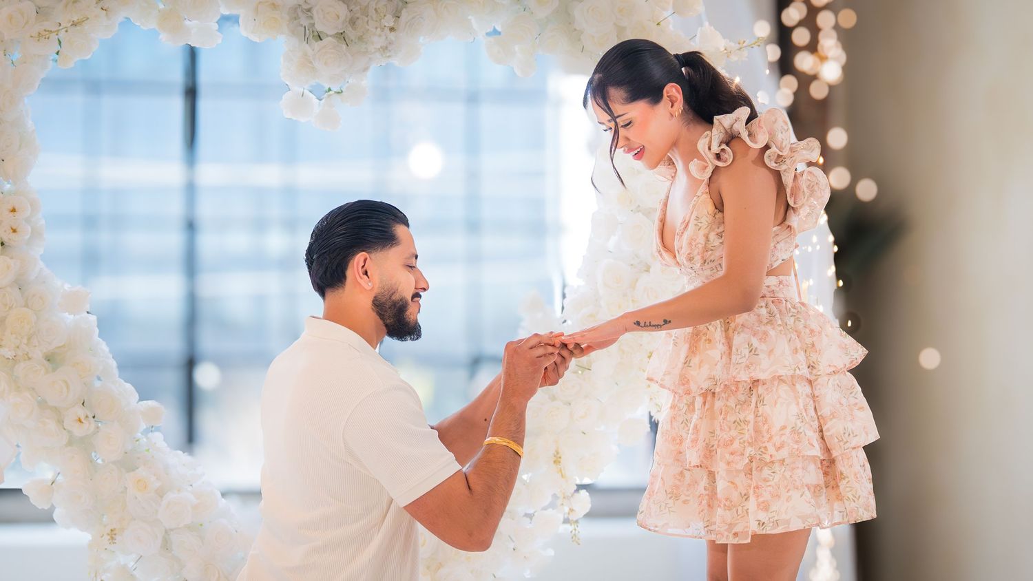 A man kneels proposing to a woman in a white dress surrounded by floral decorations.