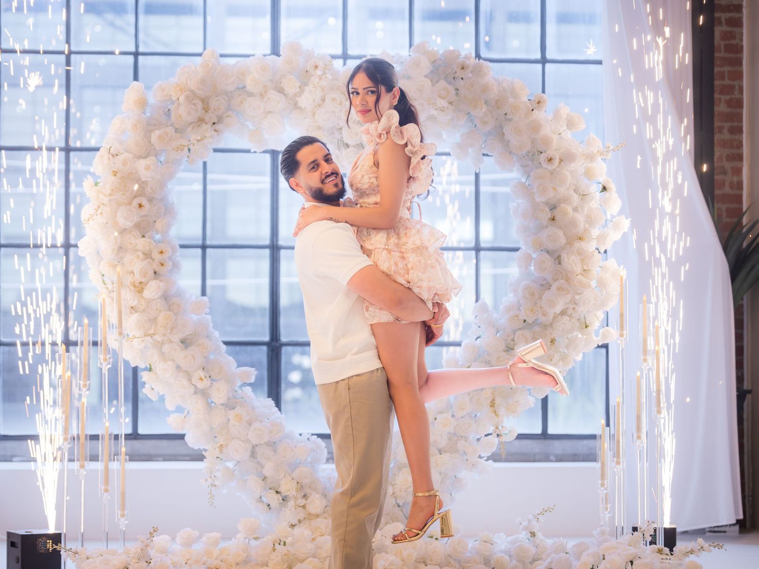 Couple celebrating engagement with man lifting woman inside white heart arch with cold sparklers.