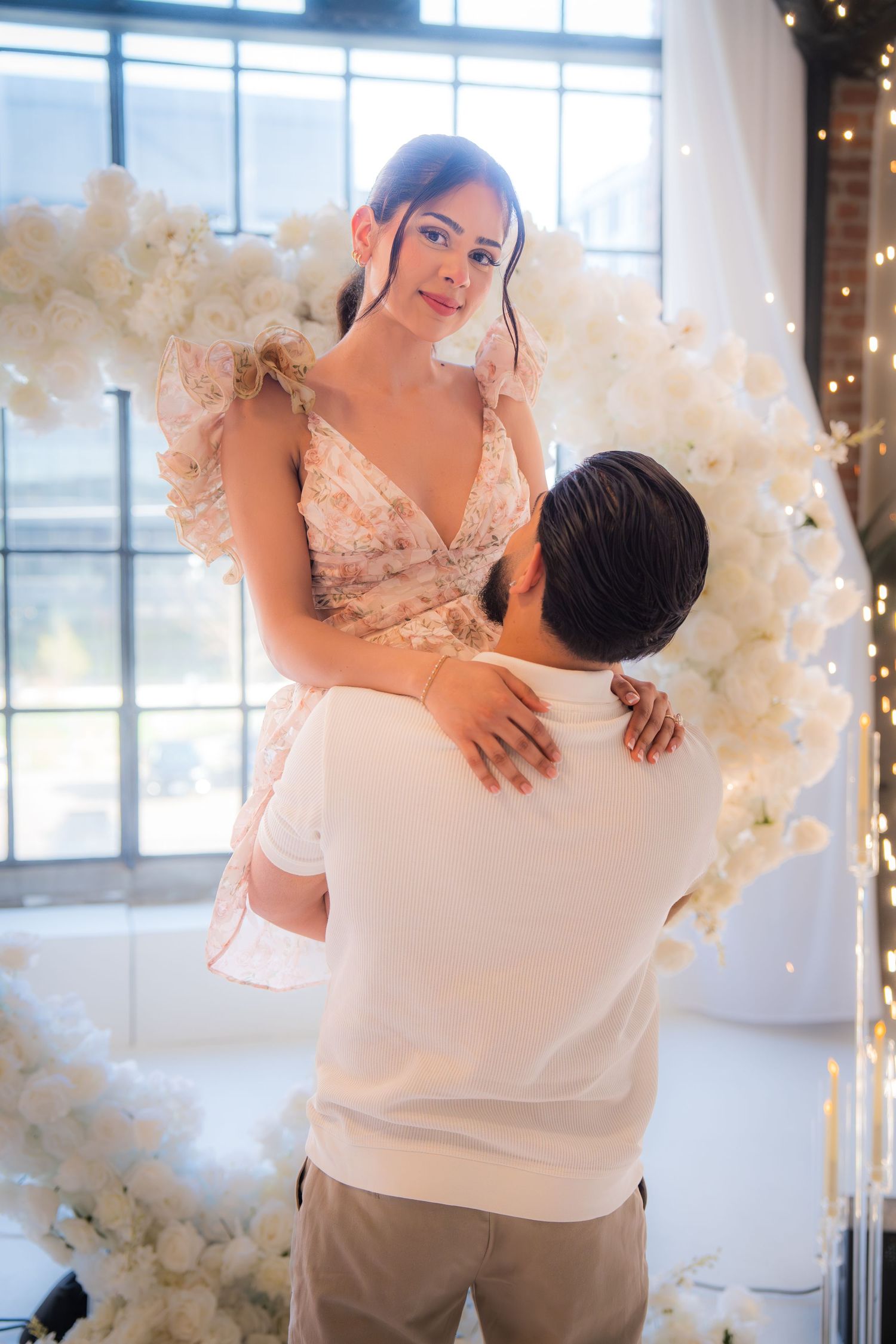 Man lifts woman in front of a white floral heart arch during a romantic proposal celebration.
