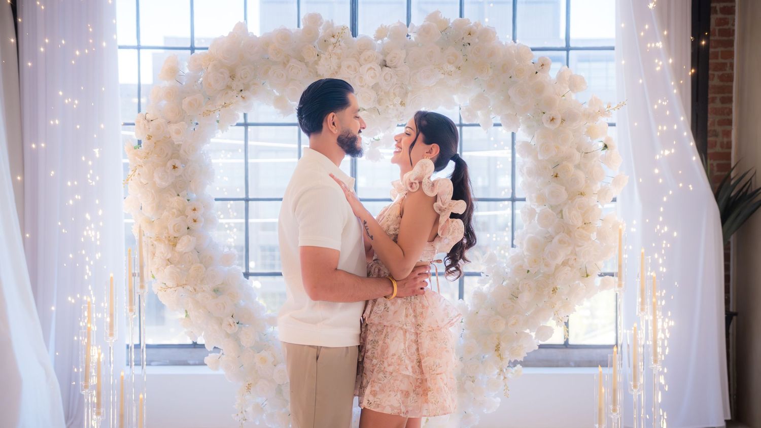 Couple sharing romantic moment under white floral heart arch with fairy lights at elegant venue.